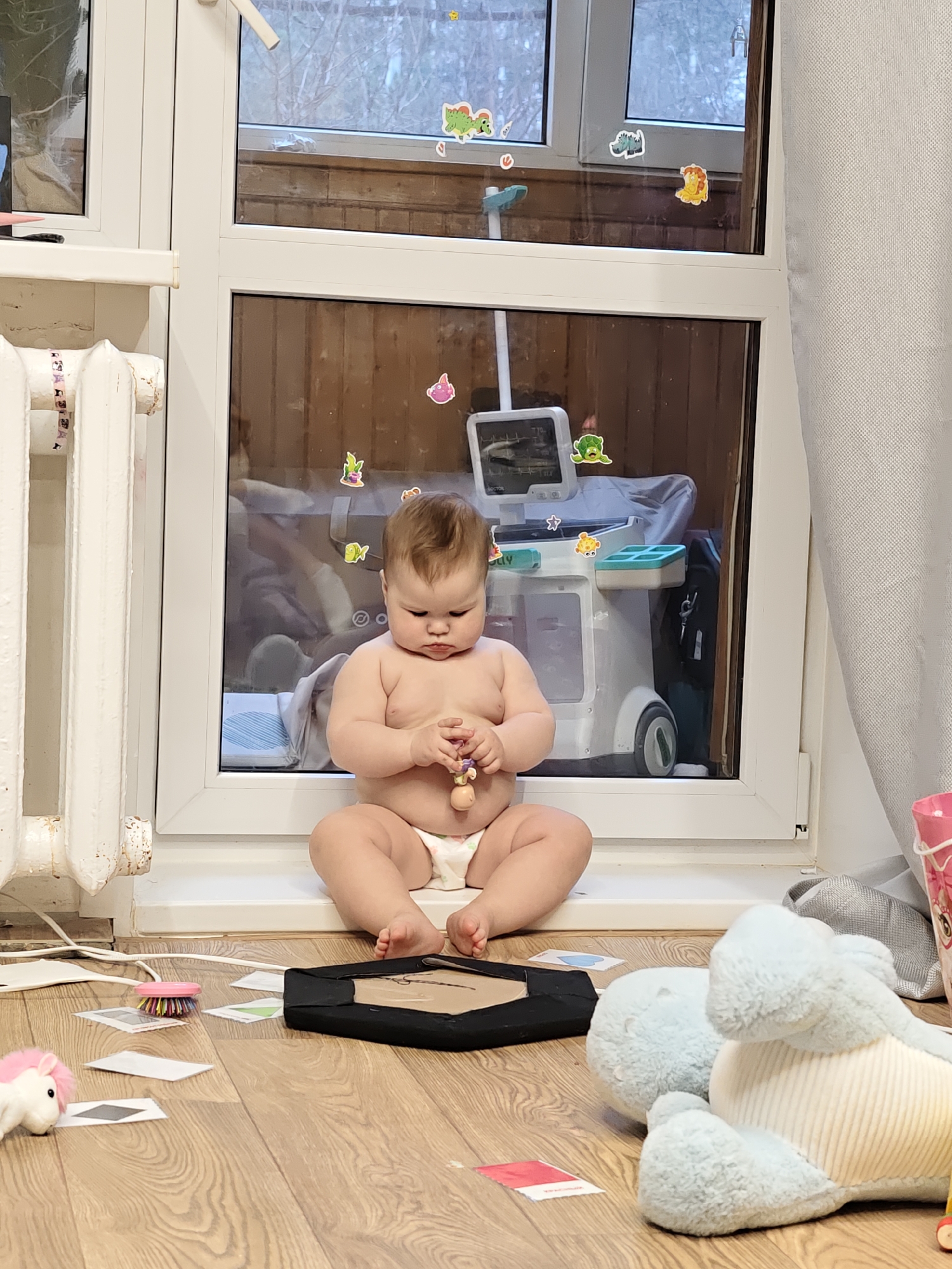 A baby wearing a diaper sits cross-legged on a white windowsill. The child holds a wooden toy. Behind the baby is a window with stickers, overlooking an outdoor scene, possibly in Chaikovsky, Russia, given the location metadata. Visible through the window is a changing table with an ultrasound machine toy. Various toys and cards are scattered on the wooden floor in front of the window, including a stuffed unicorn, a blue stuffed animal, a pink bucket, and a black square object. A white radiator is visible to the left of the window, and a grey curtain hangs to the right.