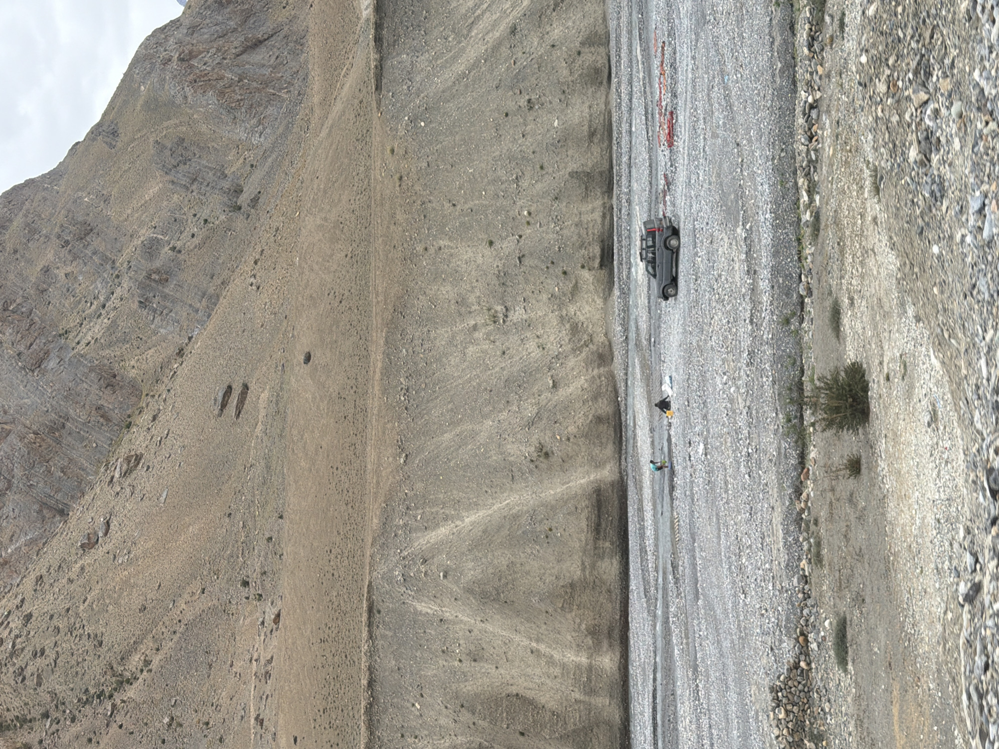 In Zangla, India, a grey SUV is parked in a rocky riverbed. Two individuals, one wearing a turquoise shirt and the other a black shirt, are in the distance, appearing to work near the river. The riverbed is covered in grey pebbles and stones. Behind the riverbed, a steep, barren hillside rises, characterized by layers of rock and soil. The sky is overcast.