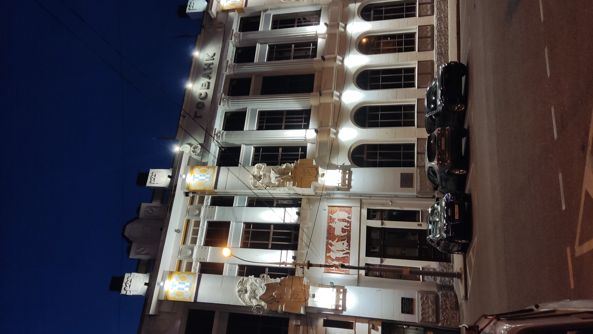 The image captures a nighttime view of a grand white building with neoclassical architectural elements, including columns and arched windows. The building is illuminated by external lights, highlighting its intricate details and a sign with the word "ГОСБАНК" in large, dark letters. Sculptural figures adorn the facade. Several cars are parked in a row along the street in front of the building. The sky above is a deep, clear blue, indicating it is nighttime. The scene appears to be in Krasnodar, Russia, given the context. There is no discernible weather activity. The overall mood is one of quiet and stately grandeur.