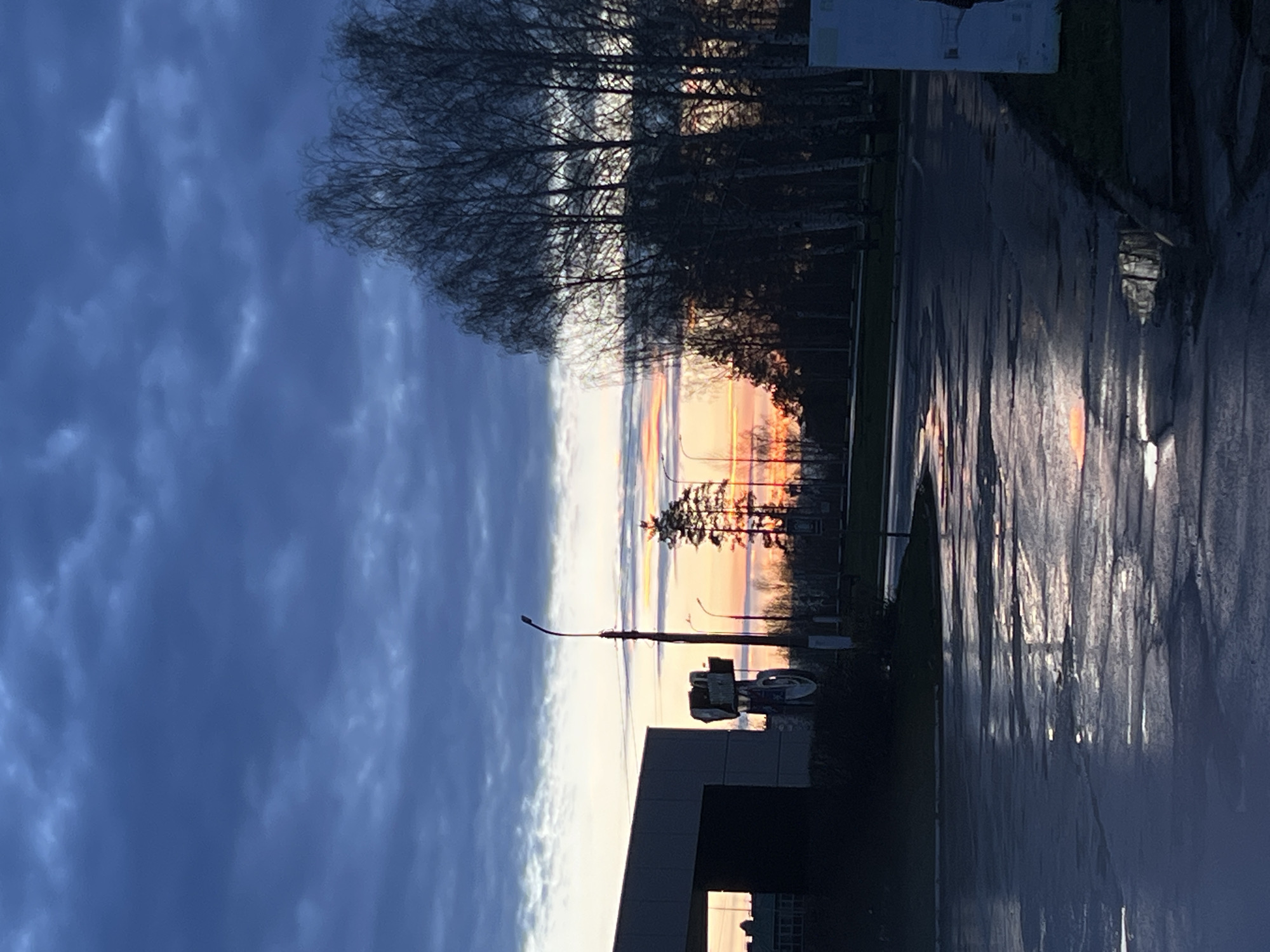 The image captures a tranquil dawn or dusk scene in Chaikovsky, Russia, dominated by a vibrant sky and reflective surfaces. The sky is split horizontally, with a broad expanse of deep blue-grey clouds on the upper left, transitioning into a brilliant display of colors along the horizon. Hues of orange, pink, yellow, and pale blue streak across the lower portion of the sky, indicative of either sunrise or sunset.
Silhouetted against this bright horizon is a line of trees, primarily bare deciduous, with some evergreen foliage interspersed. These trees border what appears to be a wet asphalt road or pathway, which occupies the foreground and reflects the dramatic sky and tree silhouettes like a mirror.
In the lower left of the frame, the dark, angular silhouette of a building is visible. A tall streetlamp with a distinctive curved arm stands in the mid-ground, along with another pole that appears to have some attached equipment or a sign, though no text is legible. The wetness of the ground suggests recent precipitation, enhancing the reflective qualities of the scene. The overall impression is one of a serene and visually striking moment at the beginning or end of the day.