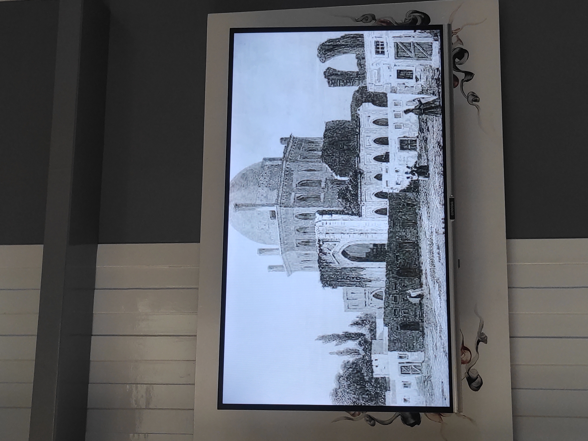 The image displays a black and white illustration of a large, domed building with surrounding structures. The illustration is shown on a television screen, which is mounted on a wall. The wall itself has a patterned design. The illustration depicts an architectural style that appears to be historical, with arches, windows, and what looks like remnants of older buildings. Several figures are visible in the foreground, dressed in traditional clothing, suggesting a historical or cultural depiction. The scene appears to be an outdoor setting with trees in the background. There is no indication of the image being located in Sumedang, Indonesia.