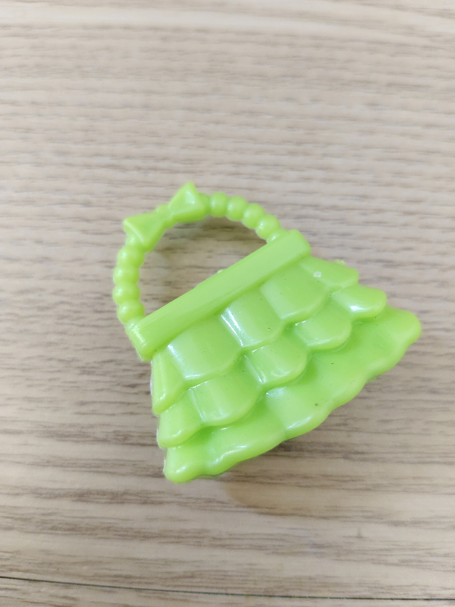 An overhead, medium shot captures a bright green plastic toy house roof with a looped handle. The roof is segmented into overlapping, scallop-like shingles and sits on a light brown wooden surface. The texture of the wood grain is visible, running horizontally across the frame. The toy is the sole subject in the image. The lighting is even, casting no significant shadows. The location is unknown.