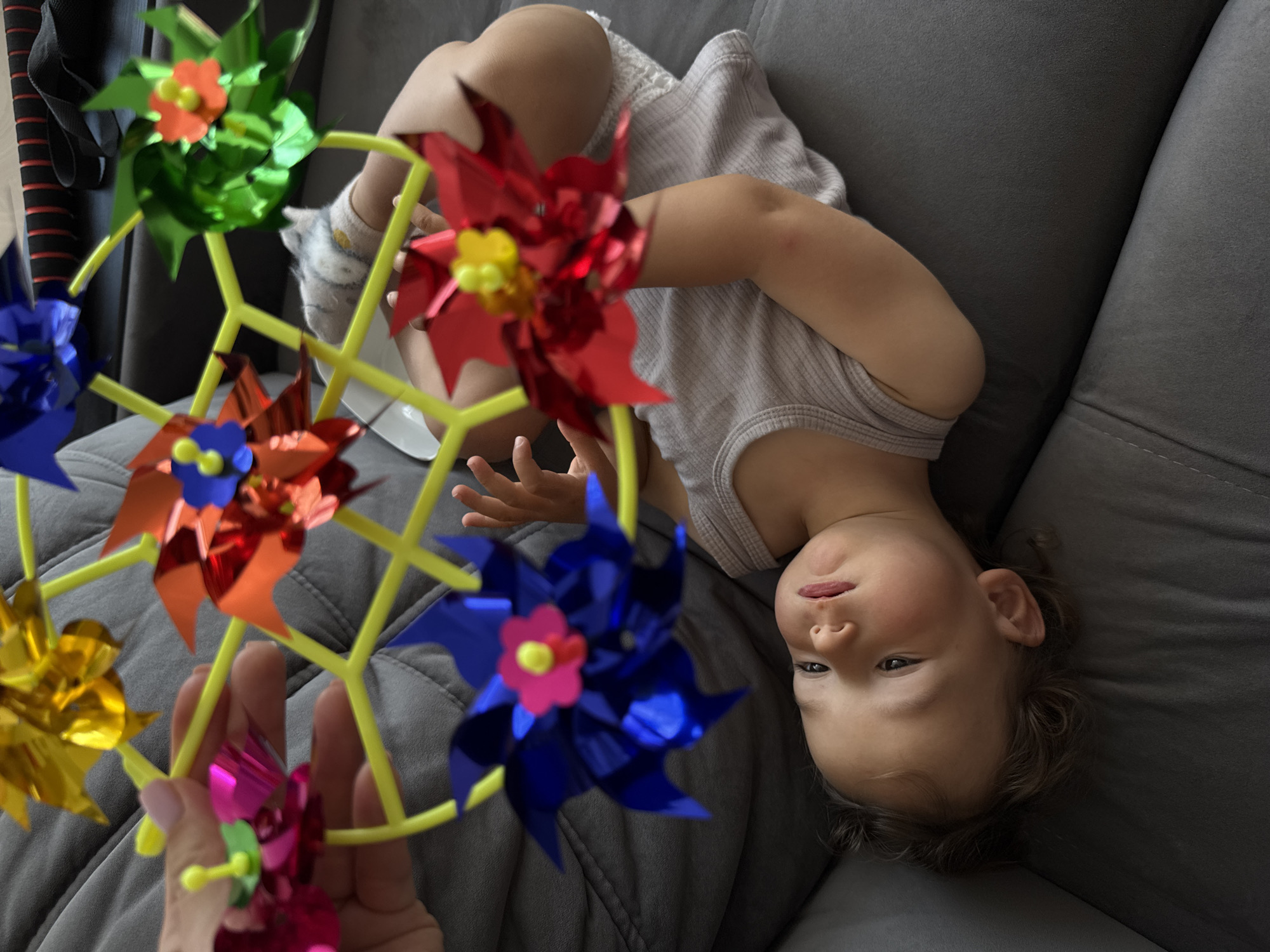 A high-angle photograph captures a young child lying supine on a grey upholstered sofa in Sochi, Russia. An adult's hand, exhibiting manicured nails with light polish, is visible in the upper left, supporting a toy mobile. This mobile consists of a yellow hexagonal plastic frame from which six small, metallic pinwheels are suspended in various colors: blue, red, green, magenta, and yellow, each with a contrasting central button. The child, dressed in a light-colored ribbed sleeveless garment, looks upwards towards the toy with a slightly open mouth. Both of the child's hands are partially raised, appearing to interact with or reach for the pinwheels. The immediate environment is composed of the textured grey sofa, with a dark, striped object partially visible in the upper background.