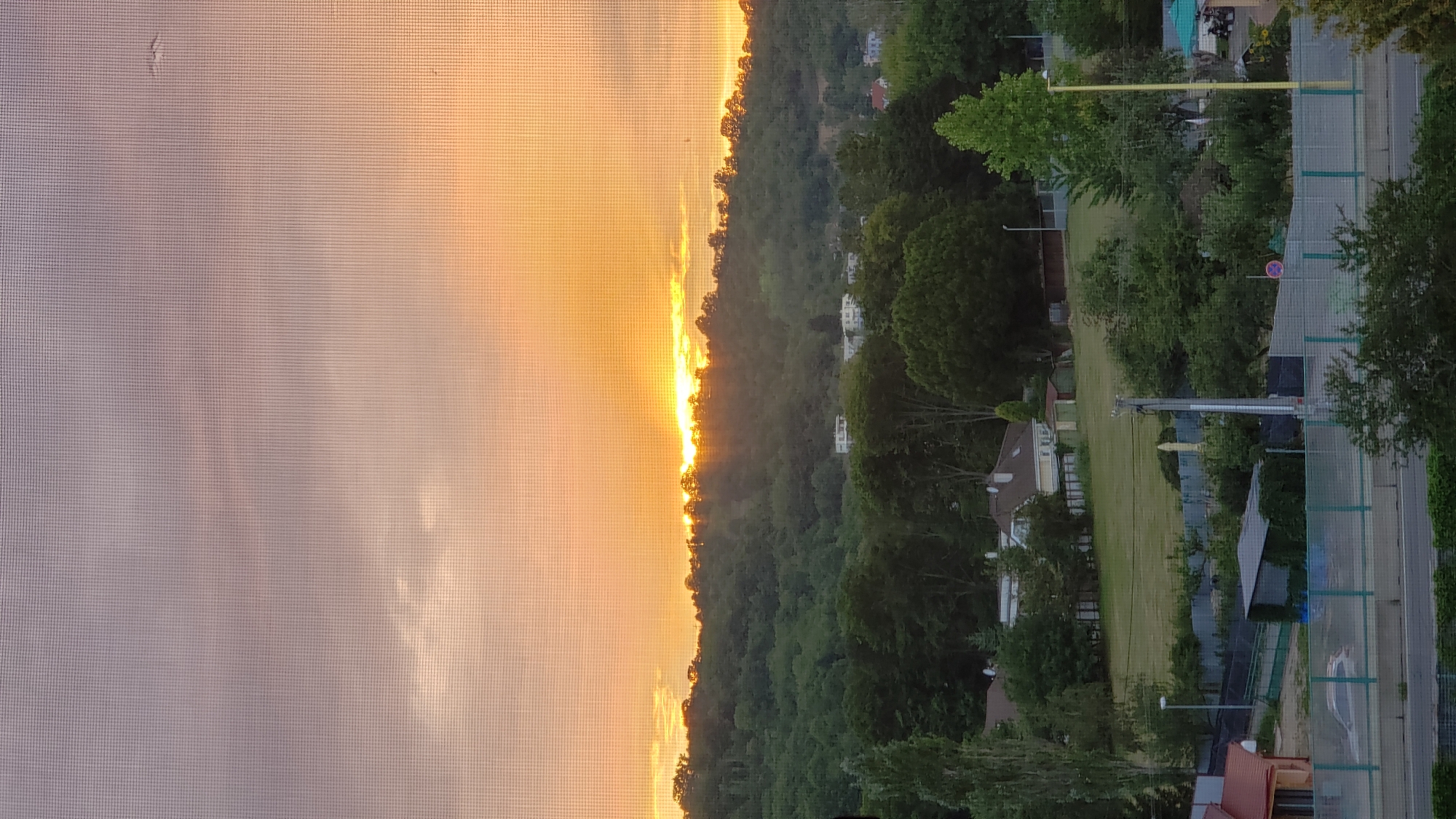 This is a photograph of a sunset over a wooded hill, likely taken from an elevated position within the city of Prešov, Slovakia. The sun is partially obscured by a treeline atop the hill, casting warm orange and yellow light across the sky and illuminating the clouds. The hill is covered in dense green trees, and several buildings are visible nestled among them and on the slopes. In the foreground, there are more trees, a grassy field, fences, and some structures that appear to be houses or sheds. A road and a car are also visible in the lower portion of the image. The overall scene depicts a natural landscape on the outskirts of a city during the golden hour.