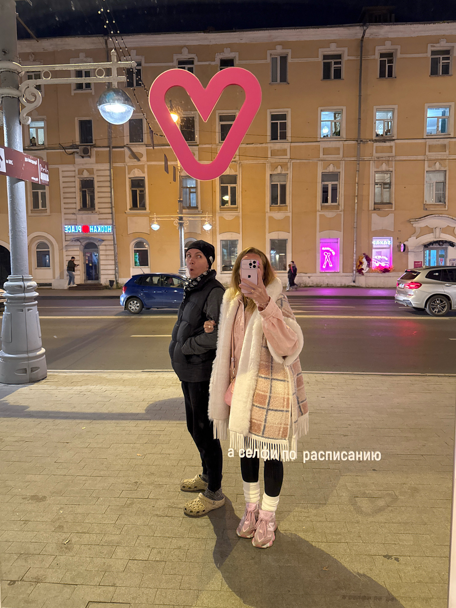 The image shows two individuals standing on a sidewalk in front of a building at night. The building is lit with warm-toned lights, and there are several windows visible. A large, pink heart-shaped decoration is positioned in the upper part of the frame, obscuring some of the building. A street lamp with a visible bulb is mounted on a pole to the left. In the background, there is a street with a blue car and a silver SUV. The sidewalk is made of concrete pavers. Text overlayed on the image reads "а селфи по расписанию" which translates to "a selfie on schedule". The individuals are facing the camera, with one taking a selfie and the other looking towards the camera. The person on the left is wearing a dark puffer vest over a long-sleeved shirt and dark pants, with a black beanie. The person on the right is wearing a plaid coat with a fluffy trim, black leggings, and pink sneakers with white socks. This scene is located in Tver, Russia, specifically on Sadovaya Street, as indicated by the signage visible in the background that reads "HOOKAH PLACE" with Russian text, and the overall urban setting.