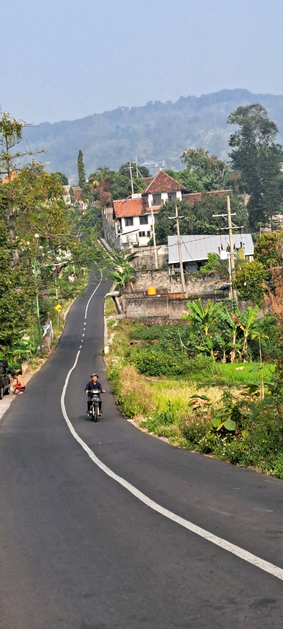 In Sukoreno, Indonesia, a motorcyclist wearing a dark shirt and a helmet rides a black motorcycle down a paved road that curves to the left. The road has a white line painted down the center. To the right of the road, there is lush green vegetation, including banana trees, and a brick wall. Further up the hill, several buildings with red-tiled roofs are visible amongst dense trees. A person can be seen on the left side of the road near a black car, partially obscured by trees. The background is dominated by a hazy mountain range.