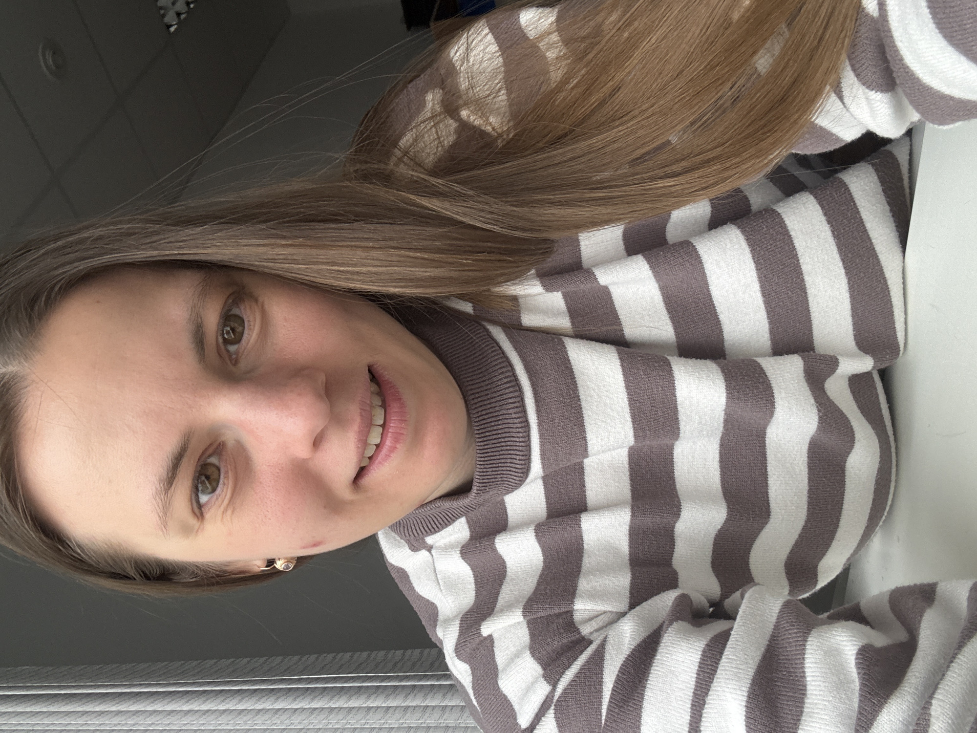 A portrait-style photograph captures a young woman smiling and looking directly at the camera in an indoor setting in Chaikovsky, Russia. She has long, straight, light brown hair that extends over her shoulders and chest. Her brown eyes convey a friendly and content expression, complemented by a gentle smile that reveals her teeth.
She is wearing a long-sleeved sweater or top featuring prominent horizontal stripes that alternate between white and a light brown or mauve hue. A small, delicate earring with a light-colored gem is visible in her right ear.
The background suggests an office or home environment. To her left, a window with horizontal blinds is partially visible, indicating natural light entering the room. Above her head, a ceiling with square panels and a circular light fixture can be seen. The right side of the background is a plain, light-colored surface, possibly a wall. No text is visible in the image.