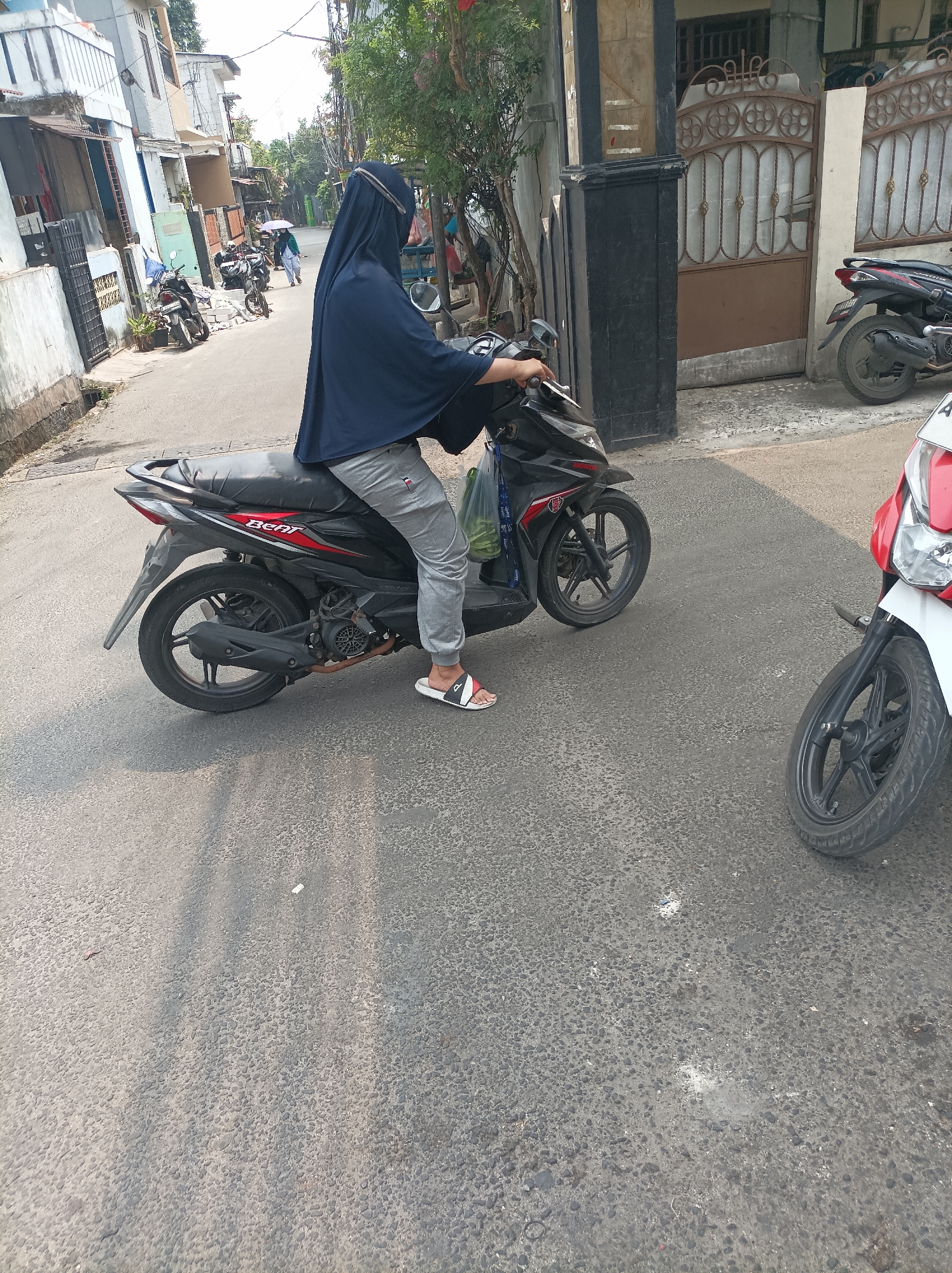 A person wearing a dark blue hijab and grey pants is sitting on a black and red motorcycle, facing to the left of the frame. They are holding the handlebars and appear to be stationary. In the background, there are buildings, other motorcycles parked, and trees. The scene is an outdoor setting, likely a street in a residential area. The pavement is grey asphalt. A red and white motorcycle is parked to the right of the frame, with its front wheel visible. The environment suggests a warm day due to the sunlight and shadows. The context implies West Jakarta, Indonesia, given the commonality of motorcycles as transportation in the region and the architectural style of the buildings.
