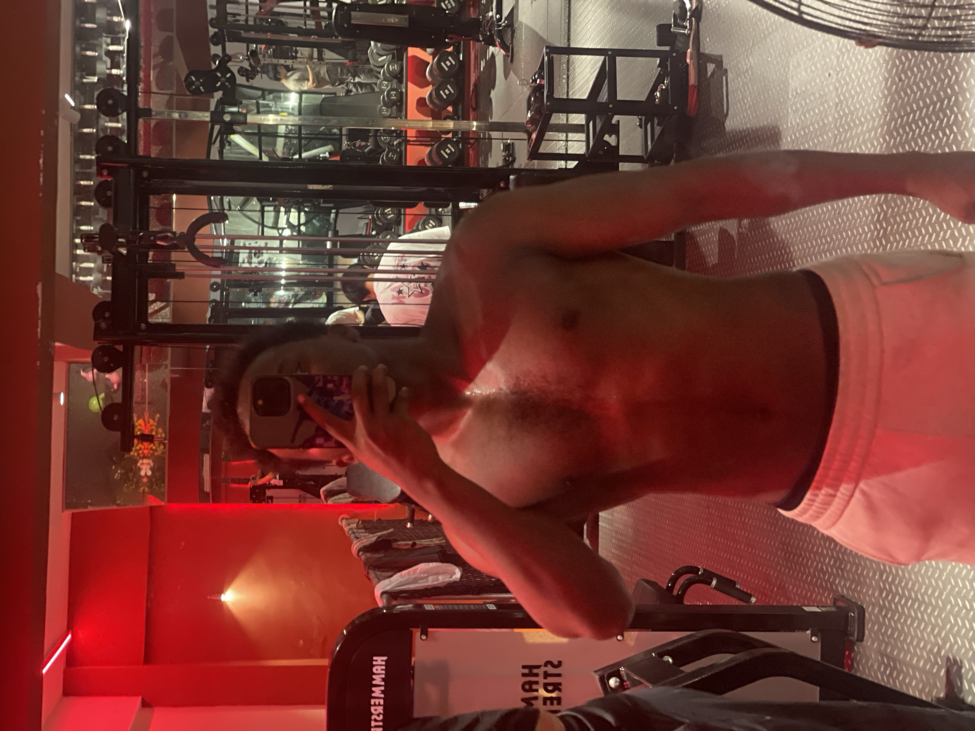 An individual is taking a mirror selfie in a gym. The individual is shirtless, wearing white sweatpants, and is holding a phone up to their face. The gym is lit with red lights, and various weightlifting equipment is visible in the background, including dumbbells and weight machines. The floor is a diamond-plate metal. The gym appears to be located in the Chongqing High-tech Zone, China.