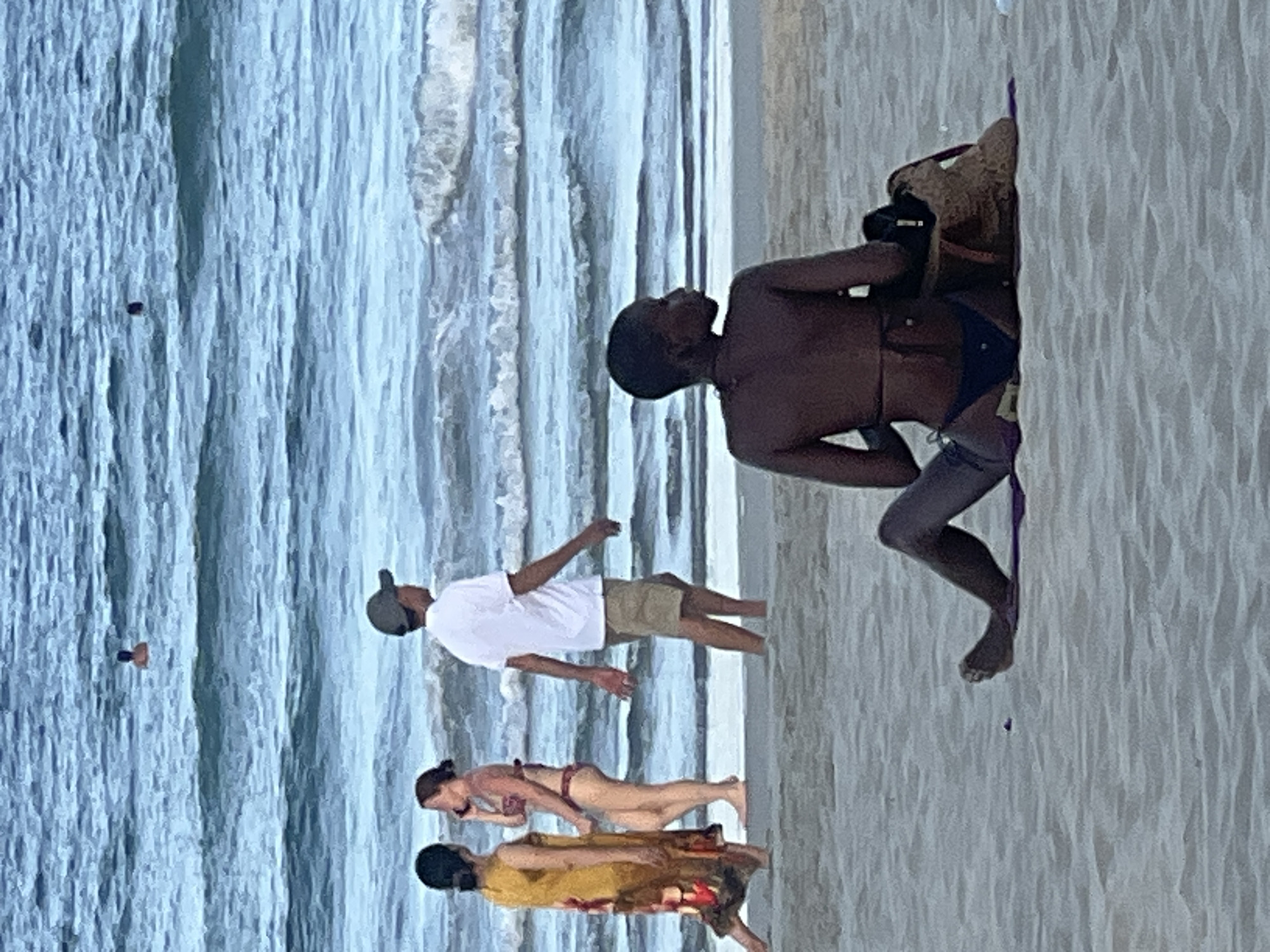 The image shows a group of people on a beach in Da Nang, Vietnam. The ocean is on the left, with gentle waves rolling in. The beach is sandy and appears to be a popular spot with several individuals present.
In the foreground, three people are walking on the sand. The person closest to the viewer is wearing a yellow dress and a black hat. Next to them is a woman in a bikini, and further back is a man in a white t-shirt and khaki shorts, also wearing a hat.
In the background, a person is seated on the sand, facing away from the camera, with a straw hat and a bag beside them. Their legs are visible, and they are wearing a dark-colored bikini bottom. Further out in the water, a person can be seen swimming.
The lighting suggests it is daytime, likely afternoon, with overcast skies. The overall mood is relaxed and casual, typical of a beach scene. There is no visible text in the image.
