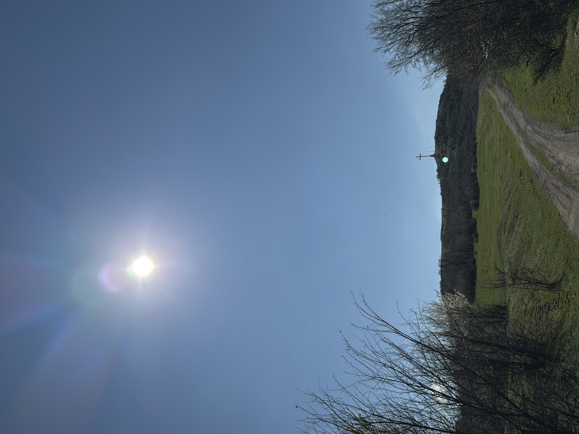 The image depicts a landscape in Bata, Bulgaria on a sunny day. The sun is visible in the left portion of the sky, accompanied by lens flares. To the right, a dirt road ascends a grassy hillside. A cross structure is present on the crest of the hill. Trees and shrubs are seen along the road and at the bottom of the frame. The sky is clear and blue.