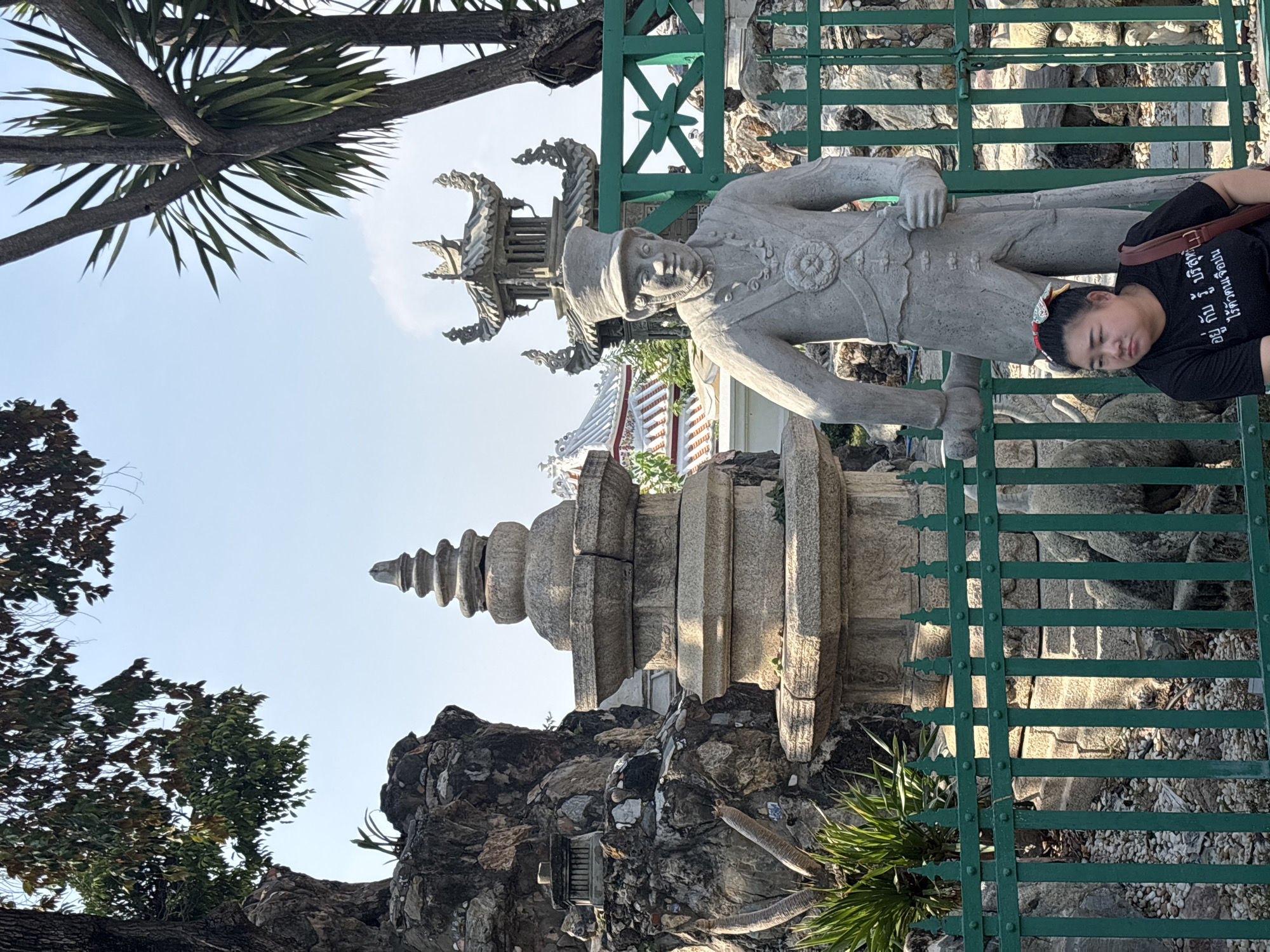 A woman rests her head on a stone statue behind a green wrought iron fence. The statue is of a European man in a military uniform and cap. Behind the statue is a pagoda or stupa structure with a series of tiers. The statue and pagoda are part of a larger, rocky structure. Some greenery and trees are visible in the background. The image was likely taken in Bangkok, Thailand, specifically at Wat Arun (Temple of Dawn), where similar statues and structures are found.