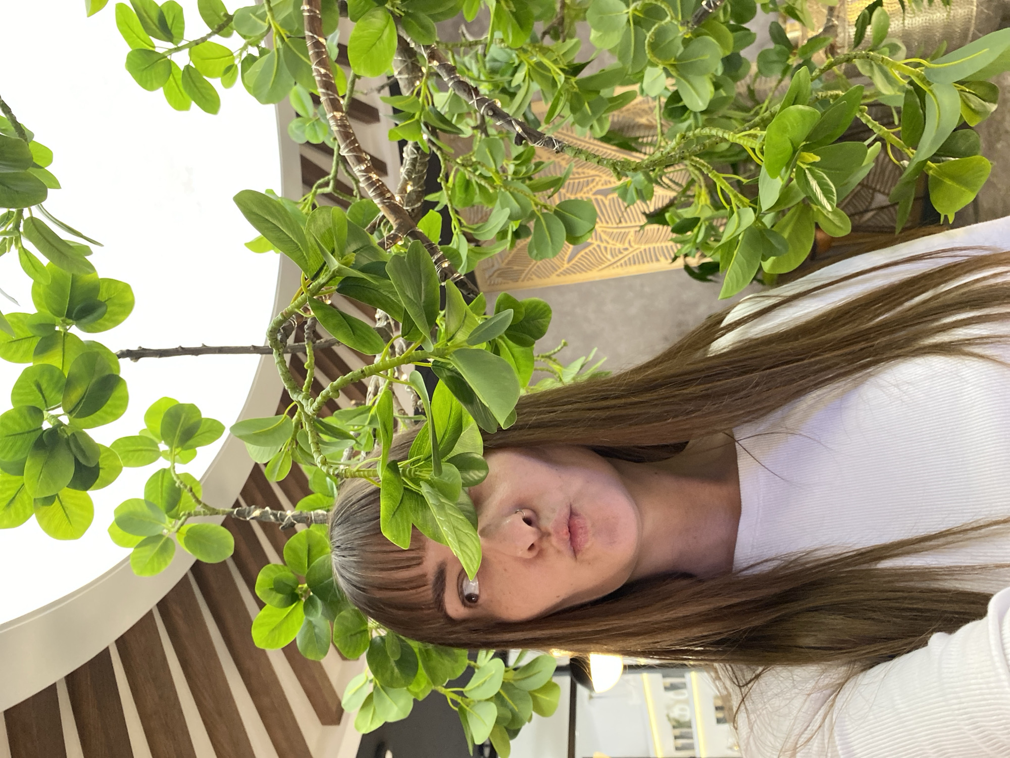 A selfie depicts a woman with light brown hair and bangs, wearing a white shirt. She is standing beneath a plant with green leaves. A small, thin silver ring is visible in her left nostril. The background features a wall with a patterned design and wooden slats arranged in a circular pattern. Based on visual characteristics, no definitive evidence suggests this image was taken in Votkinsk, Russia.