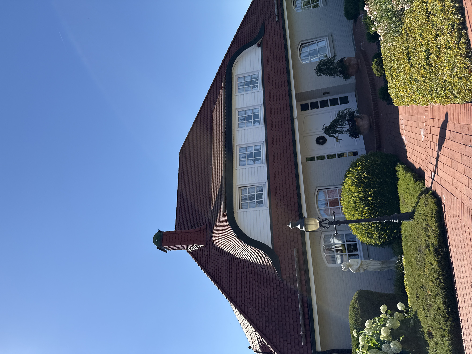 The image shows a large, two-story house with a distinctive red-tiled roof and a dormer window. The house is painted a light gray color. The front of the house features a white double door with a wreath, flanked by windows. Potted plants are situated on either side of the entrance. To the left of the house, there is a garden area with neatly trimmed bushes, a white statue, and a large white hydrangea bush in bloom. A brick pathway leads to the front door. A black lamppost stands in the foreground. The sky is clear and blue. The house appears to be located in a residential area. There are no people or animals visible in the image. The image depicts a property on a sunny day.