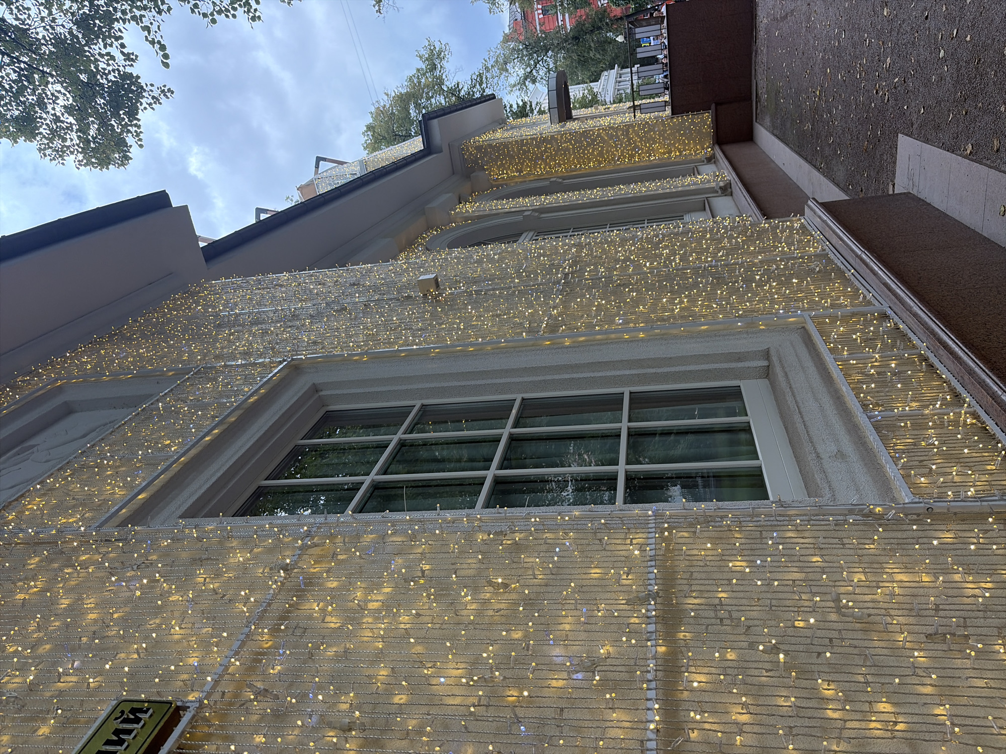 The image shows the facade of a building in Moscow, Russia, adorned with numerous strings of warm white lights. The lights are arranged vertically, creating a curtain-like effect across the building's exterior. A large, multi-paned window is visible on the left side of the image, reflecting the surrounding environment. The building has a light-colored textured wall and a darker base. In the background, trees with green foliage and a cloudy sky are visible. Further down the facade, arched windows and architectural details can be observed. A sidewalk with scattered leaves is in the foreground, and part of a dark red wall or structure is present to the right. The overall scene suggests a decorated building, possibly for an event or season, in an urban setting.