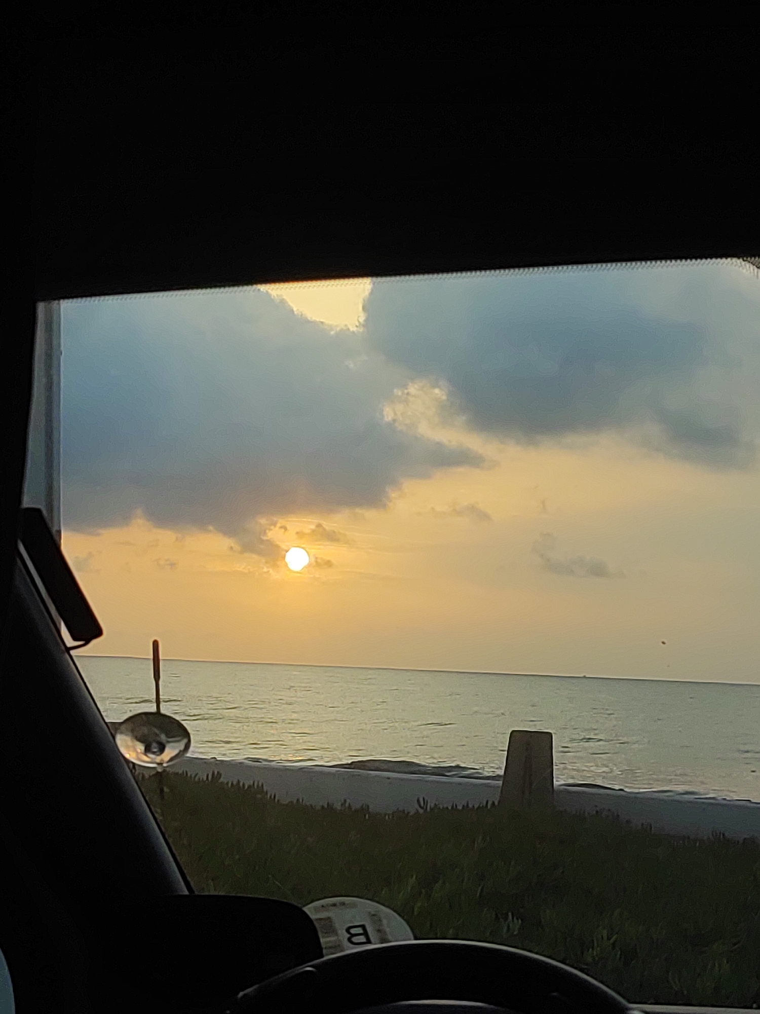 The image depicts a sunset over the ocean, viewed through the windshield of a vehicle. The sun, a bright white disc, is positioned slightly left of the center of the frame, casting a warm, golden light across the sky and the water. Dark gray clouds are scattered across the upper portion of the sky, contrasting with the lighter, more diffused clouds near the horizon.
The ocean stretches out to the horizon, appearing calm with gentle waves. Along the shoreline in the foreground, there is a strip of green vegetation, likely beach grass or low-lying shrubs. A dark, rectangular concrete structure stands on the beach, to the right of the center. On the left side of the image, a circular mirror is suspended from a thin rod, reflecting the scene. The lower part of the image shows the interior of the vehicle, including a portion of the steering wheel and what appears to be a circular sticker with the letter "B" on it. The overall lighting suggests it is dusk or early evening. The location is described as Playa, Spain.