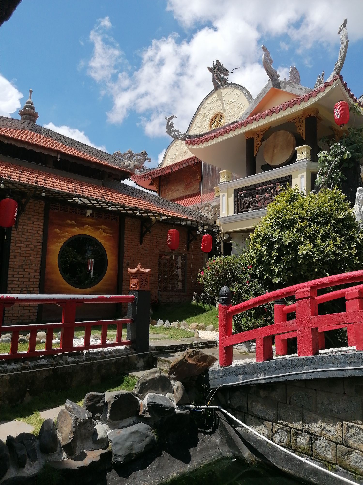 This image captures a serene Buddhist temple nestled in the city of Lâm Viên - Đà Lạt, Vietnam. The architecture features traditional red-tiled roofs, ornate eaves, and decorative figurines adorning the rooftops. A prominent red bridge with intricate carvings leads into the temple grounds, where lush greenery and carefully placed rocks create a tranquil atmosphere. Red lanterns hang from the eaves, adding a touch of vibrant color to the scene. A large drum is visible within one of the temple's structures, suggesting a space for meditation and spiritual practices. The sky above is a bright blue, dotted with fluffy white clouds, indicative of a pleasant day.