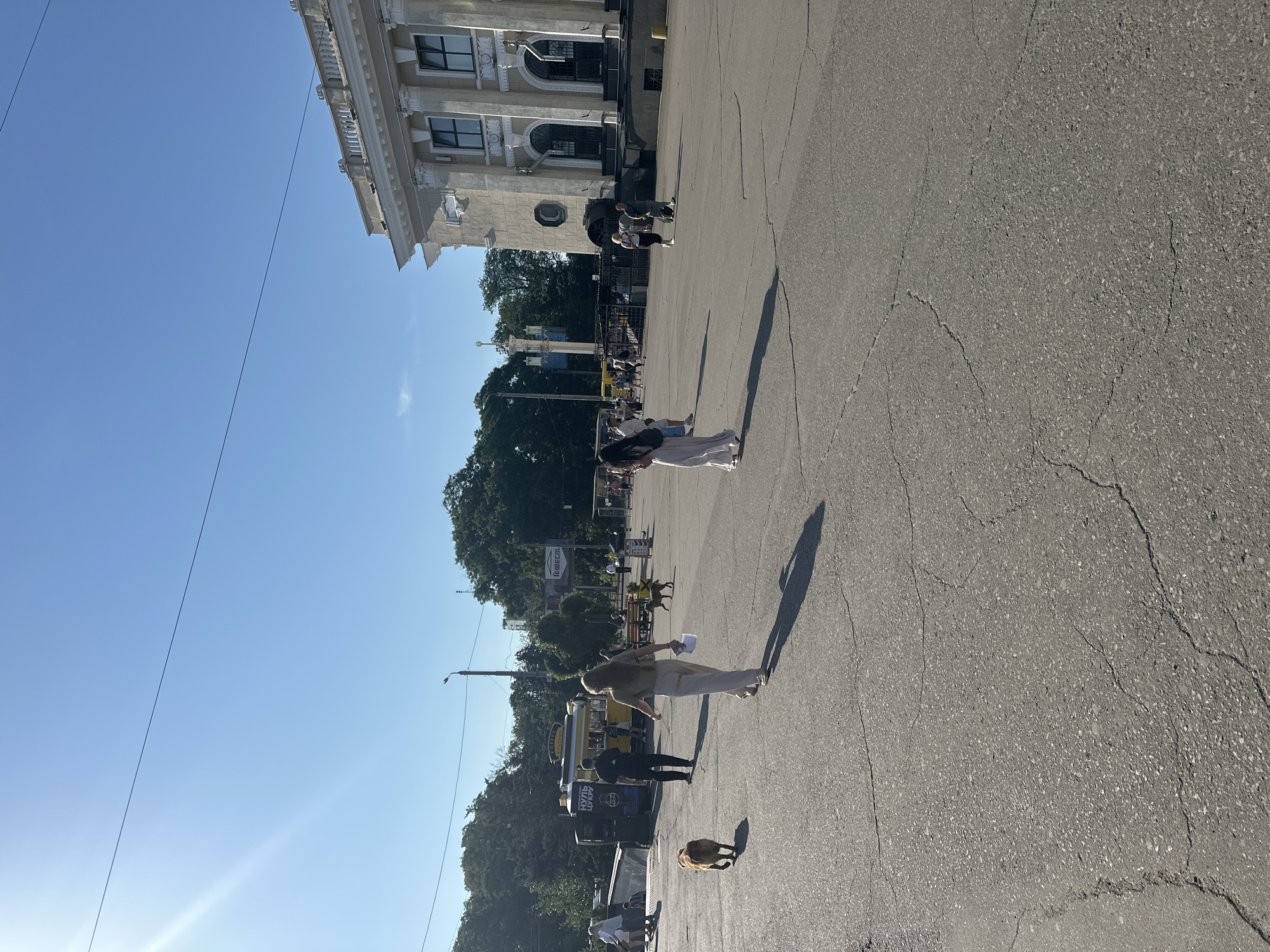 The image depicts an outdoor scene in Odesa, Ukraine, under a clear blue sky with bright sunlight casting long shadows. In the foreground, a cracked asphalt surface is visible. A medium-sized, tan-colored dog is walking across the pavement towards the left side of the frame. Behind the dog, a woman with long blonde hair, wearing a light-colored top and pants, walks away from the camera. She is holding a piece of paper in her right hand.
In the middle ground, several people are present. To the left, a food truck with signage including "COFFEE STATION" and what appears to be Cyrillic text is visible. A man in a black t-shirt and dark pants is walking away from the food truck. Further back, a person is walking a dog, and other individuals are seated on benches or walking. Trees line the background, and a large, ornate building with classical architectural elements, including columns and arched windows, is situated on the right side of the frame. The building appears to be a significant structure, possibly a public institution. There are also utility poles and wires visible. The overall atmosphere suggests a public gathering space on a sunny day.
