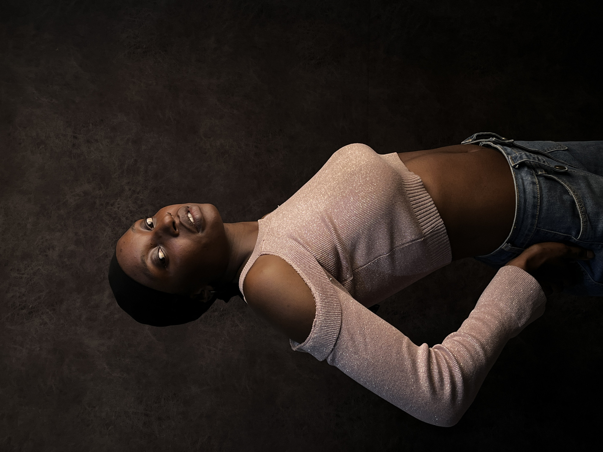 A dark-skinned woman is lying on her back against a dark, textured background. She is wearing a light pink, glittery, cropped long-sleeve shirt and blue jeans. She is looking up with a slight smile. Her head is covered with a dark cap. No location, including Barakin Akawo, Nigeria, is discernible.