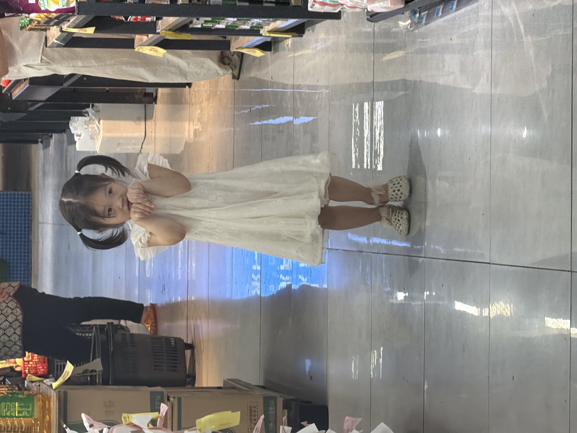 In a brightly lit interior, possibly a supermarket aisle in Jiangjin, China, a young East Asian girl stands centrally. She has dark hair styled in two pigtails. She is wearing a white lace dress and embellished sandals. Her hands are clasped together in front of her face, and she appears to be looking directly at the camera. The floor is tiled and highly reflective, mirroring the overhead lights and the environment. To her left, a dark plastic shopping basket is visible, and further back, a person in dark pants and orange footwear is partially visible. To her right, shelves stocked with products are present, and another person in light-colored pants is standing. The overall impression is of a candid moment captured within a commercial setting.