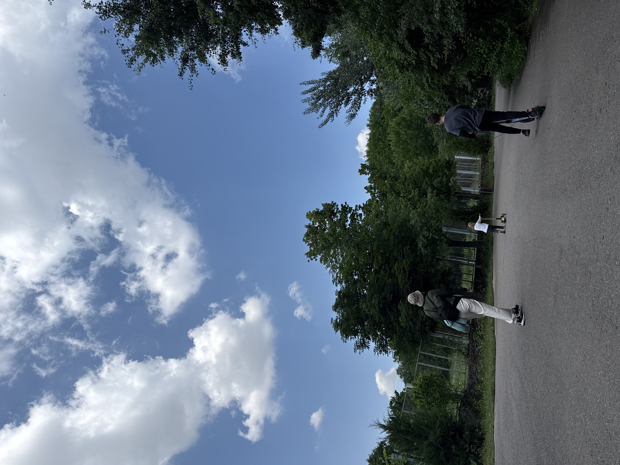 On a paved pathway in Tashkent, Uzbekistan, there are three individuals. One person is riding a scooter, another appears to be walking with a device on the ground, and the third is also walking. Lush green trees line the pathway, and a clear blue sky with scattered white clouds is visible above. A fence runs along the side of the path.