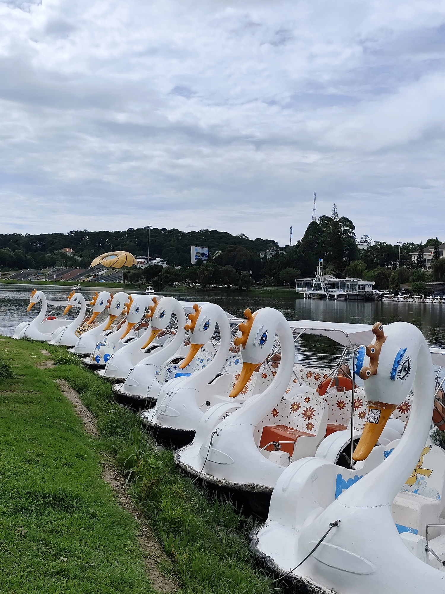 The image depicts a row of white swan-shaped paddle boats lined up along the grassy bank of a lake. The boats are primarily white with orange beaks and blue eye details. Their seats feature a brown and white floral pattern. The lake stretches out to the horizon, with visible structures and foliage on the far shore. Notable landmarks visible across the lake include a yellow saucer-shaped building and a white building with an observation tower. The sky is overcast with gray clouds. This location is in Da Lat, Vietnam.