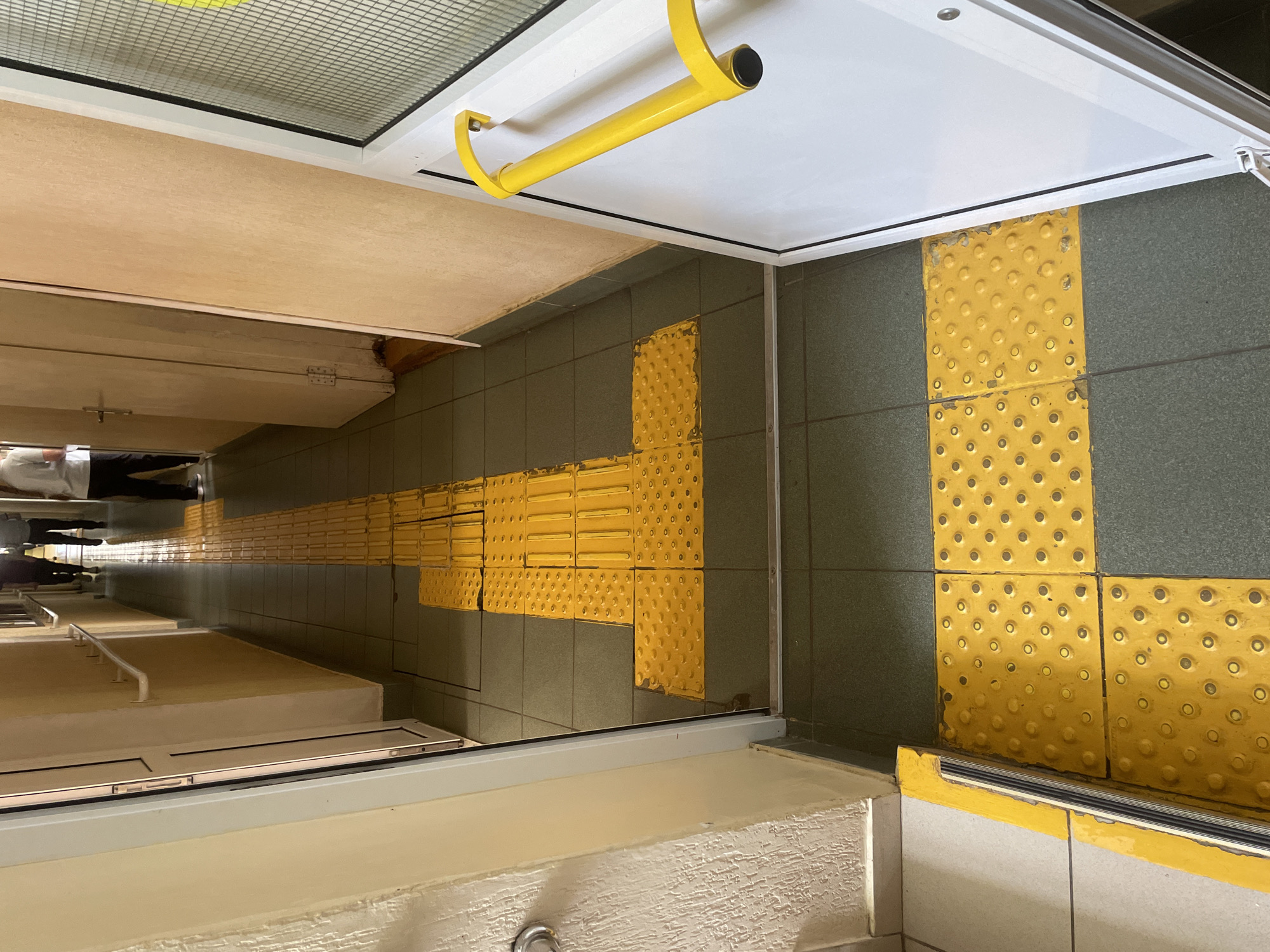 The image depicts the interior of what appears to be a subway or train station, potentially in Chaikovsky, Russia. The predominant features are the tactile paving (textured ground surface indicators) in bright yellow, contrasting with the grey-green tiled floor. These tactile tiles are arranged in strips and T-junctions, likely to guide visually impaired individuals. Part of a train car is visible with yellow handrail. A few people can be seen in the far distance. The overall environment is clean.