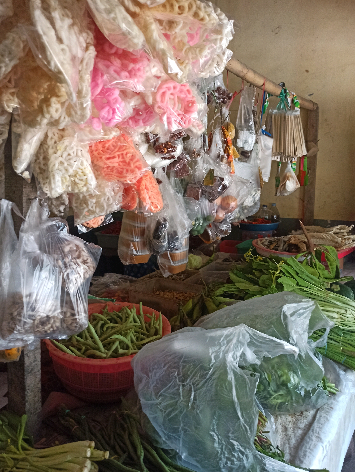 This image depicts a market stall in Tlogosari, Indonesia, featuring an array of food items and dry goods. The foreground is dominated by fresh vegetables, including bundles of long beans and leafy greens, some of which are packaged in transparent plastic. Behind these, a red woven basket overflows with green beans. Hanging from a bamboo rack above are various pre-packaged snacks and ingredients. These include colorful ring-shaped crackers in shades of white, pink, and orange, as well as other items wrapped in clear plastic, some appearing to be dried food or snacks. Further back, several blue and red bowls are visible, containing what might be dried fish or chilies. The background shows a wall and a table, suggesting an indoor or semi-covered market setting. The lighting is diffused, and the overall impression is of a busy, functional market stall. No people are visible in the frame.