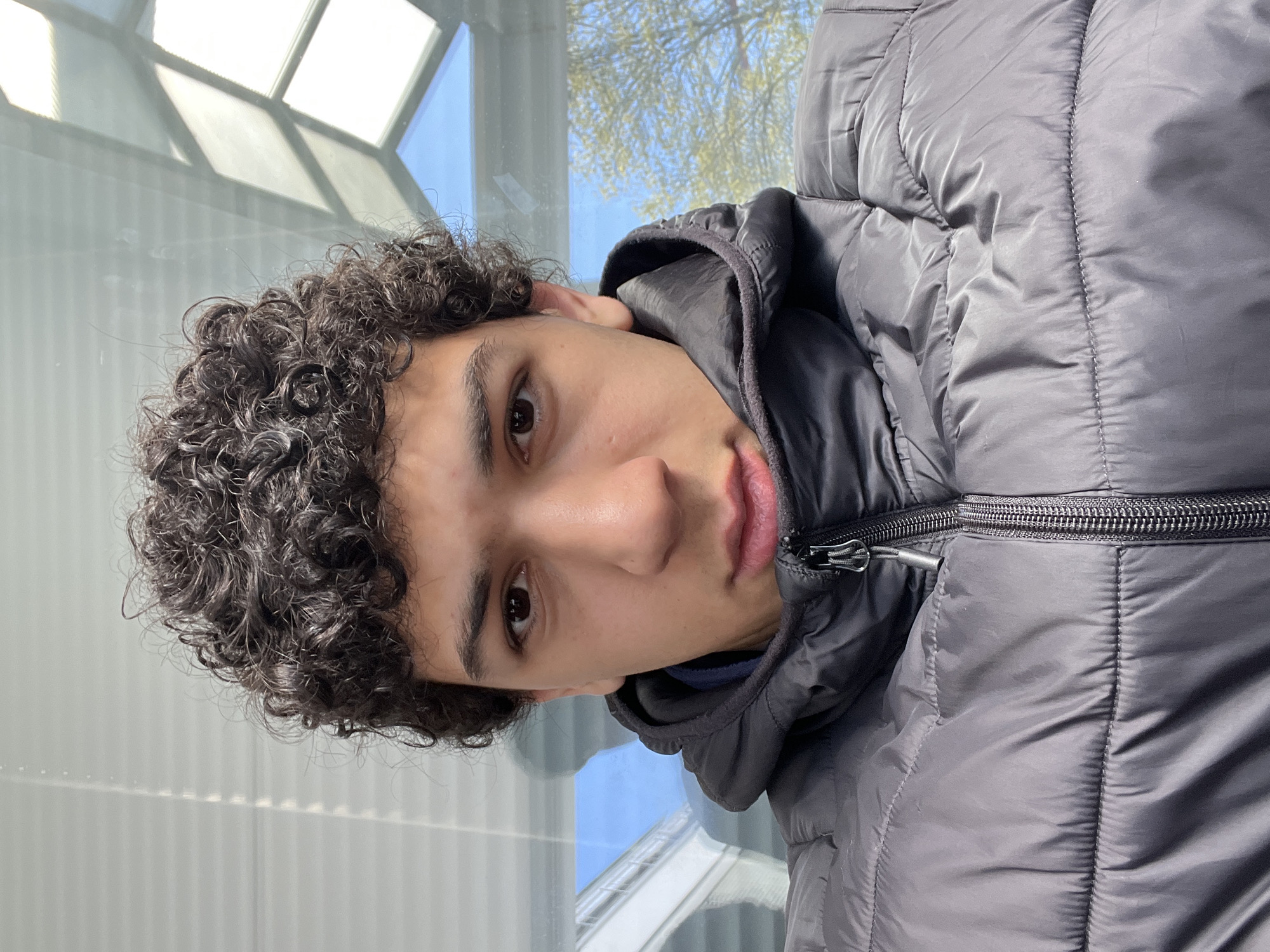A young man with curly dark hair, wearing a black puffer jacket, is pictured close-up. The background shows a window with a geometric pattern and a view of a tree with sparse leaves against a blue sky. Based on the architectural style and available context, it is possible this photo was taken in the city of Schiltigheim, France, although precise location confirmation cannot be determined solely from the image.