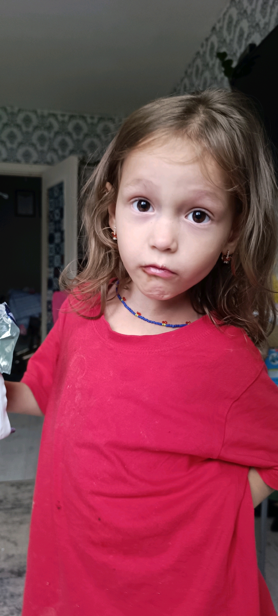 A young girl with light brown hair and brown eyes is shown in a medium shot, looking directly at the camera. She is wearing a red t-shirt and a blue beaded necklace with red and yellow beads. Her lips are slightly pursed. The background is blurred, showing a patterned wallpaper and a doorway. The lighting suggests an indoor setting, possibly a home. The image does not contain any specific landmarks or context to determine its location within the city of Chaikovsky, Russia.