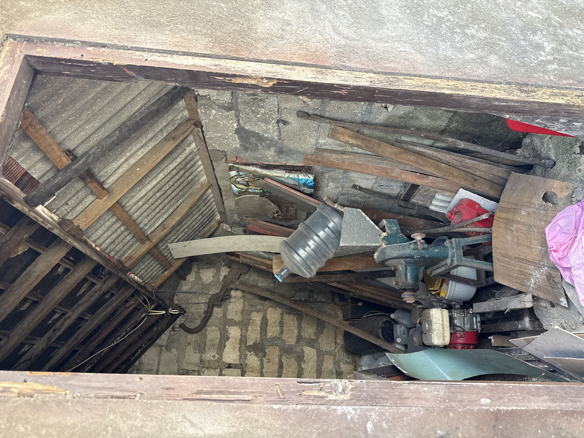 The image depicts a cluttered storage area viewed from above, likely within a building in Pejukutan, Indonesia. The space is filled with construction debris, including wooden planks, corrugated metal roofing, concrete blocks, and other unidentified materials. A water jug is visible, along with a small engine and various containers. The environment appears to be poorly maintained and possibly abandoned. No people are visible in the image.