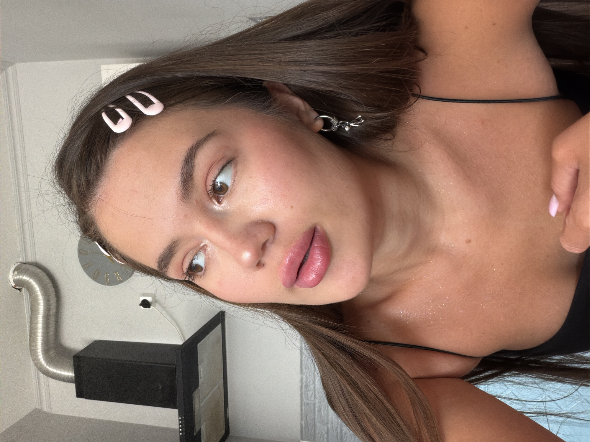 The image shows a young woman with long brown hair and light skin, wearing a black spaghetti strap top. She has two pink barrettes in her hair and a silver earring with a dangling charm. She is looking off to the side with her lips slightly parted. Her skin appears to have a subtle shimmer, possibly from glitter or sweat. In the background, there is a kitchen hood with a metal vent pipe and a clock on the wall. The location is not specified as being in the city of Sirius, Russia.