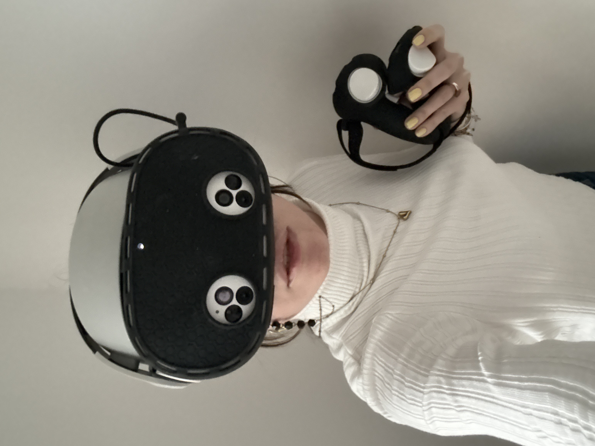 A person wearing a white turtleneck sweater is seen from a low angle, holding a black and white VR headset in their left hand and a black controller with white buttons in their right hand. The headset has a black faceplate with three camera lenses on each side. The controller has a black strap and two white buttons. The person has yellow nail polish on their fingernails. A gold necklace is visible around their neck. The background is a plain white wall. It is not possible to determine the location of the photo as Moscow, Russia.