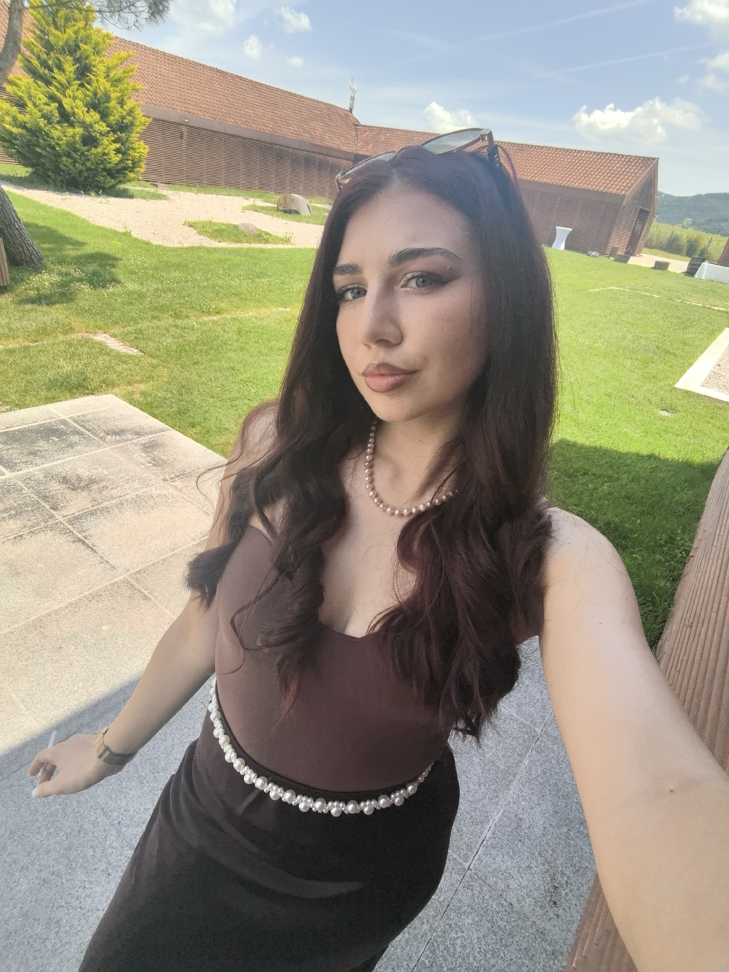 The image depicts a young woman posing for a selfie in an outdoor setting in Mogilovo, Bulgaria, on a clear, sunny day. The woman, positioned in the foreground, has long, dark brown, wavy hair, light-colored eyes, and light makeup including defined eyebrows and nude-toned lipstick. She wears sunglasses perched on her head. Her attire consists of a dark brown or maroon sleeveless top and a long, dark skirt, cinched at the waist with a belt made of white pearls. A necklace of light pinkish pearls adorns her neck. Her left hand is visible, with white-painted nails, holding a thin white object, possibly a cigarette or vape pen, between her fingers. A gold-toned watch with a brown strap is on her left wrist.
She stands on light-colored tiled or stone paving. Beyond her, a well-maintained green lawn extends towards the background. In the middle ground, a vibrant yellow-green conical shrub and parts of a tree trunk are visible on the left, along with patches of light-colored gravel or sand and a large grey rock. The background features several buildings with distinct terracotta tile roofs and horizontal wooden paneling, appearing to be single-story structures with some architectural angles. Two white pedestal-like objects are visible on the grass near the buildings on the right. The sky is bright blue with scattered white cumulus clouds, and distant green hills are visible on the horizon to the right. A wooden slatted barrier runs along the right edge of the frame.