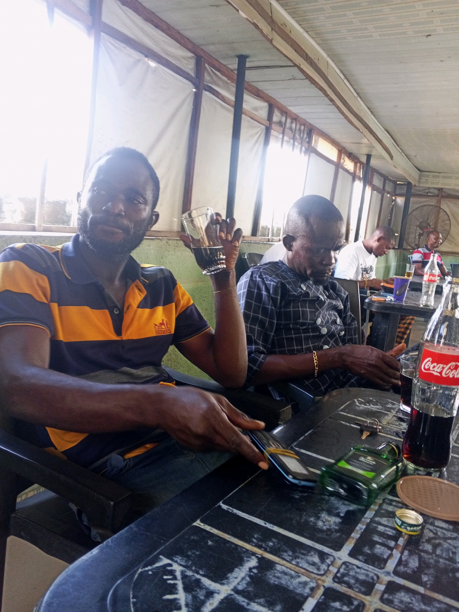 Two men are seated at a table outdoors, likely in a restaurant or bar in Zango-ndaji, Nigeria. The man in the foreground, wearing a blue and yellow striped polo shirt, holds a glass of dark liquid. He has a beard and is looking towards the camera. To his right, another man in a dark, checkered shirt with white buttons is seated, looking down at his phone or hands. He wears a gold bracelet. On the table in front of them are various items, including a bottle of Coca-Cola, a glass of dark liquid, a mobile phone, keys, and a small green bottle. In the background, other individuals are visible, and a fan is mounted on the wall. The setting appears to be a covered outdoor area with a corrugated metal roof and white fabric or tarp covering the sides. The lighting suggests it is daytime, with strong light coming from the left.