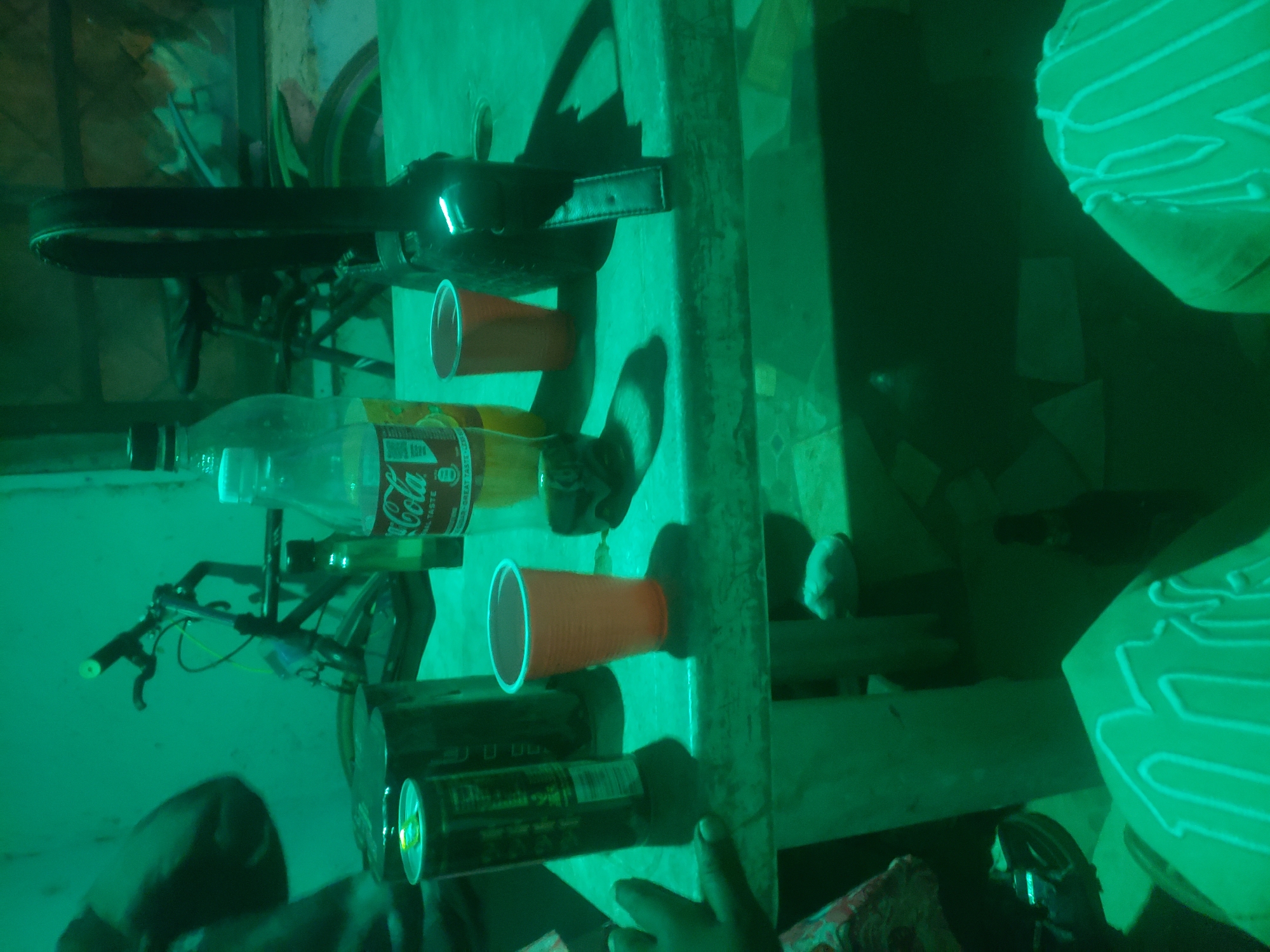 The image shows a tabletop with various objects, including a Coca-Cola bottle, two red cups, a can of Monster energy drink, and a bicycle tire lock. A bicycle is partially visible in the background. The location is Unknown, Unknown. Two individuals are partially visible. One is wearing a patterned shirt.