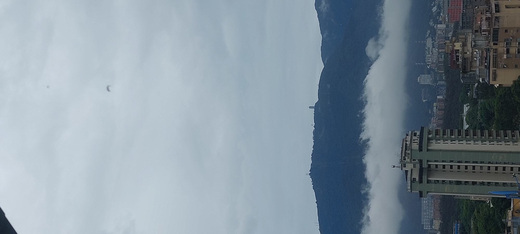 The image is an outdoor shot of a city landscape taken on an overcast day. A tall, dark, tree-covered mountain runs vertically through the center of the image. Buildings of different heights and colors are visible on the right side of the mountain. White clouds are present on both sides of the mountain, obscuring some of the buildings. The sky is predominantly gray, covering the left side of the image. A few indistinct dark spots are scattered in the sky. The city is likely Caracas, Venezuela.