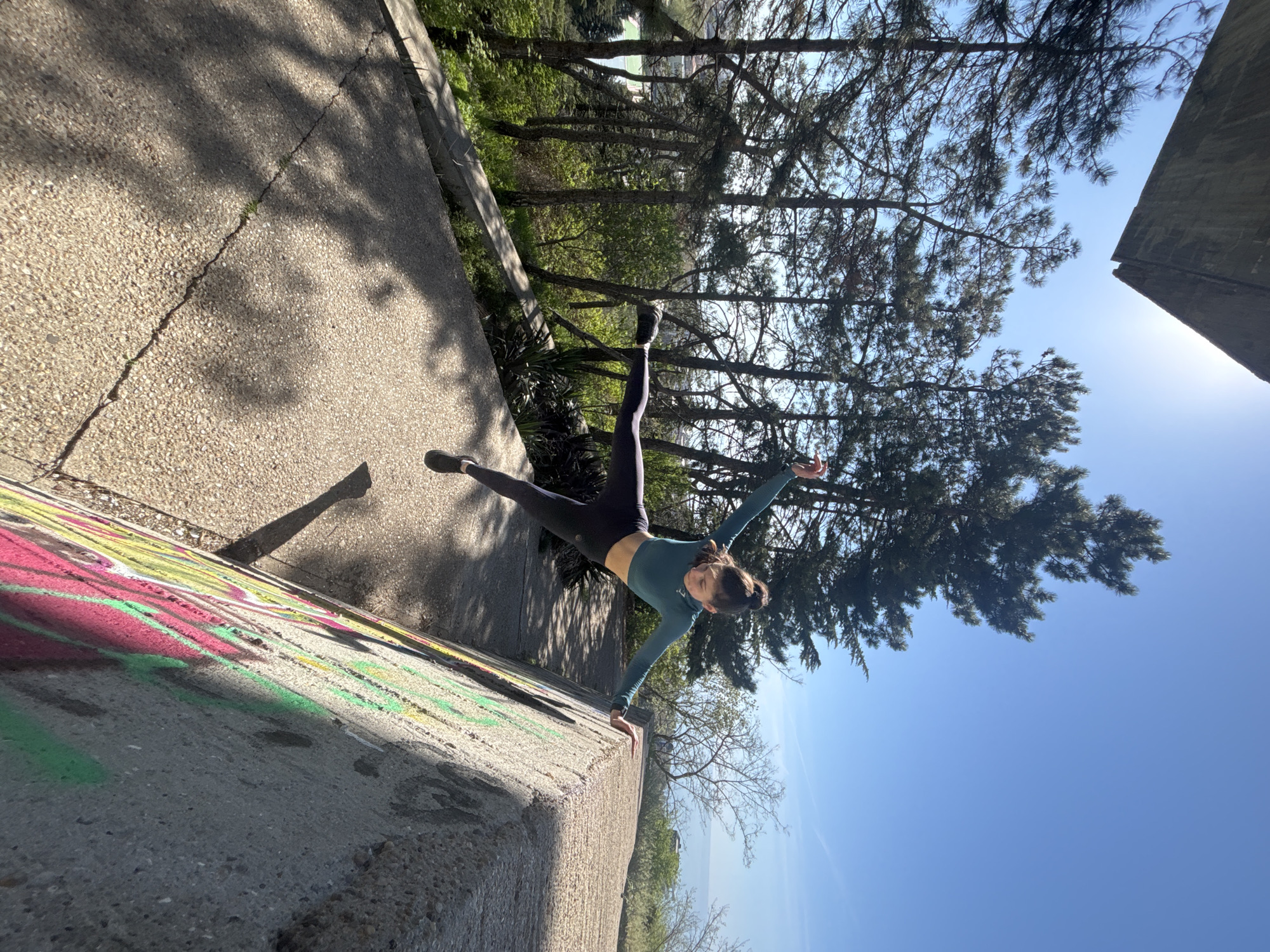 A woman is doing a handstand on a concrete wall in an outdoor, urban environment. The wall is marked with colorful graffiti. The woman is wearing a long-sleeved teal top and dark leggings. Trees are visible in the background, along with a clear blue sky. The location appears to be a city park or urban area.