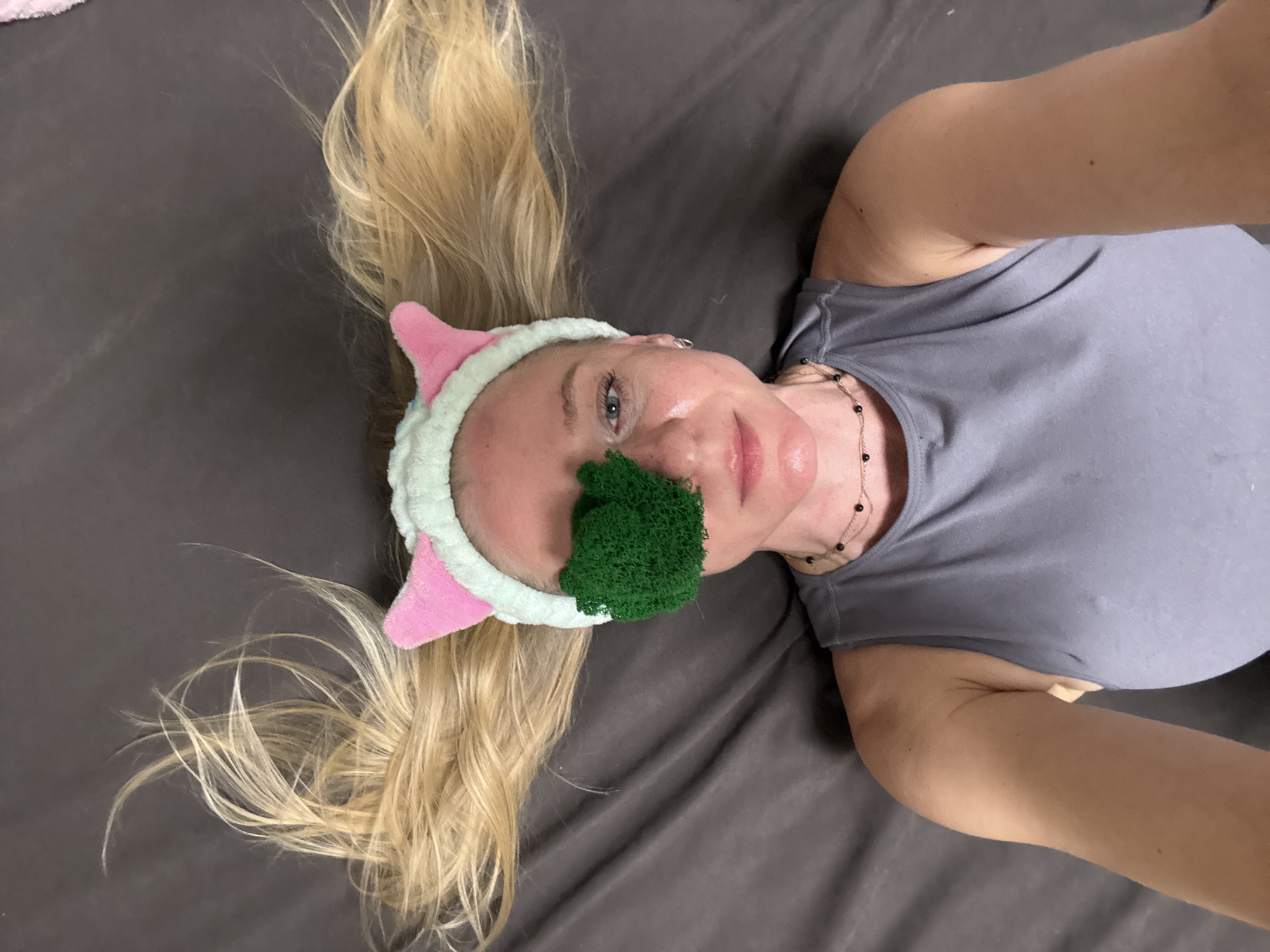 A woman lies on a grey surface, possibly a bed or floor, with blonde hair splayed around her head. She wears a grey tank top and a plush headband with pink cat ears. A green, textured object covers part of her face, possibly a facial mask or sponge. The location is unknown.