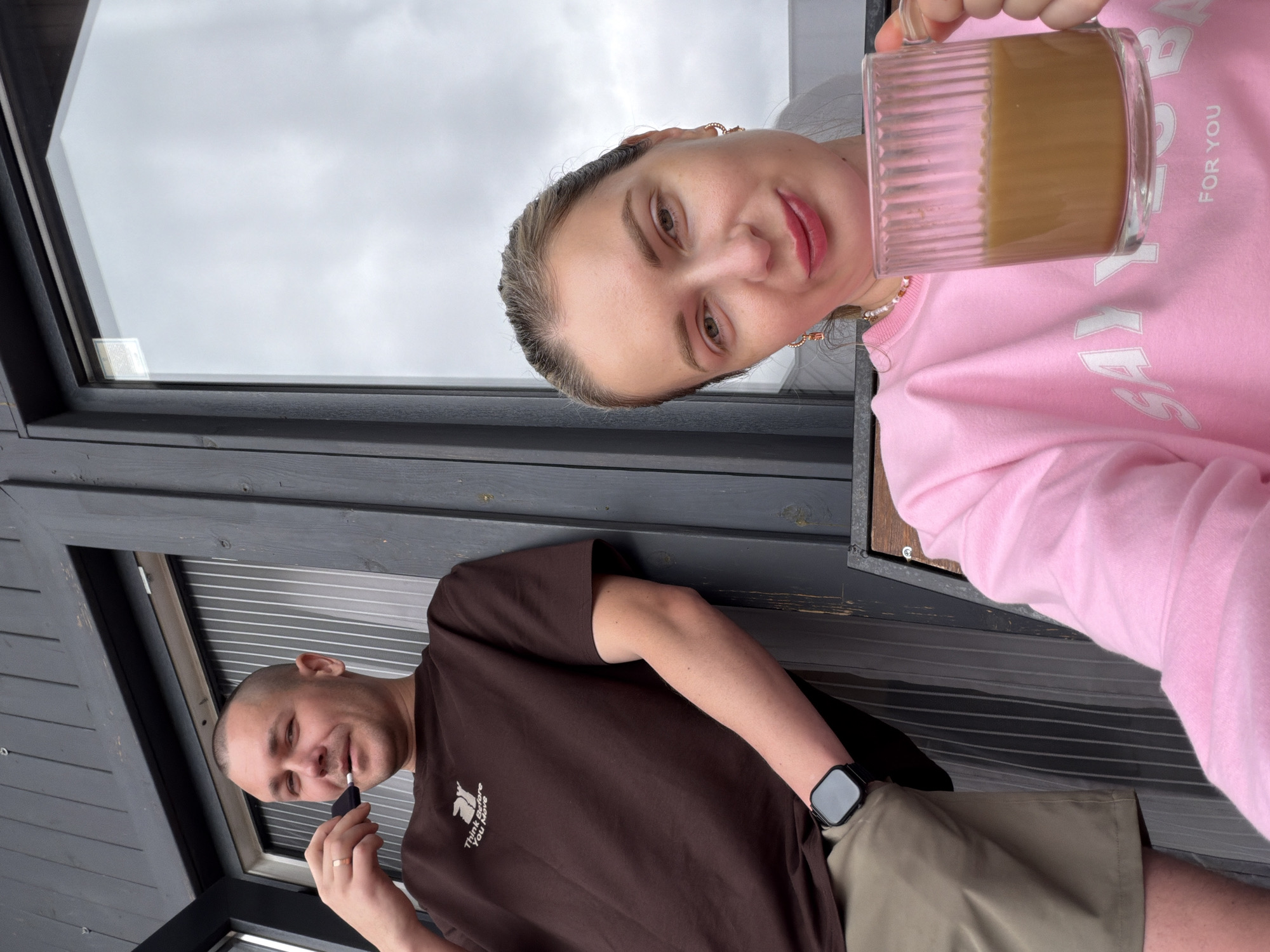 In Семилуки, Russia, a man with a shaved head, wearing a brown t-shirt and khaki shorts, stands leaning against a dark gray wooden wall. He has a ring on his left hand and is holding a dark object to his mouth. Next to him, a woman with blonde hair pulled back, wearing a pink sweatshirt and a necklace, holds a clear glass mug filled with a light brown beverage. The background consists of a large window with a cloudy sky visible through it and a slatted partition. The scene appears to be outdoors, possibly on a porch or balcony.
