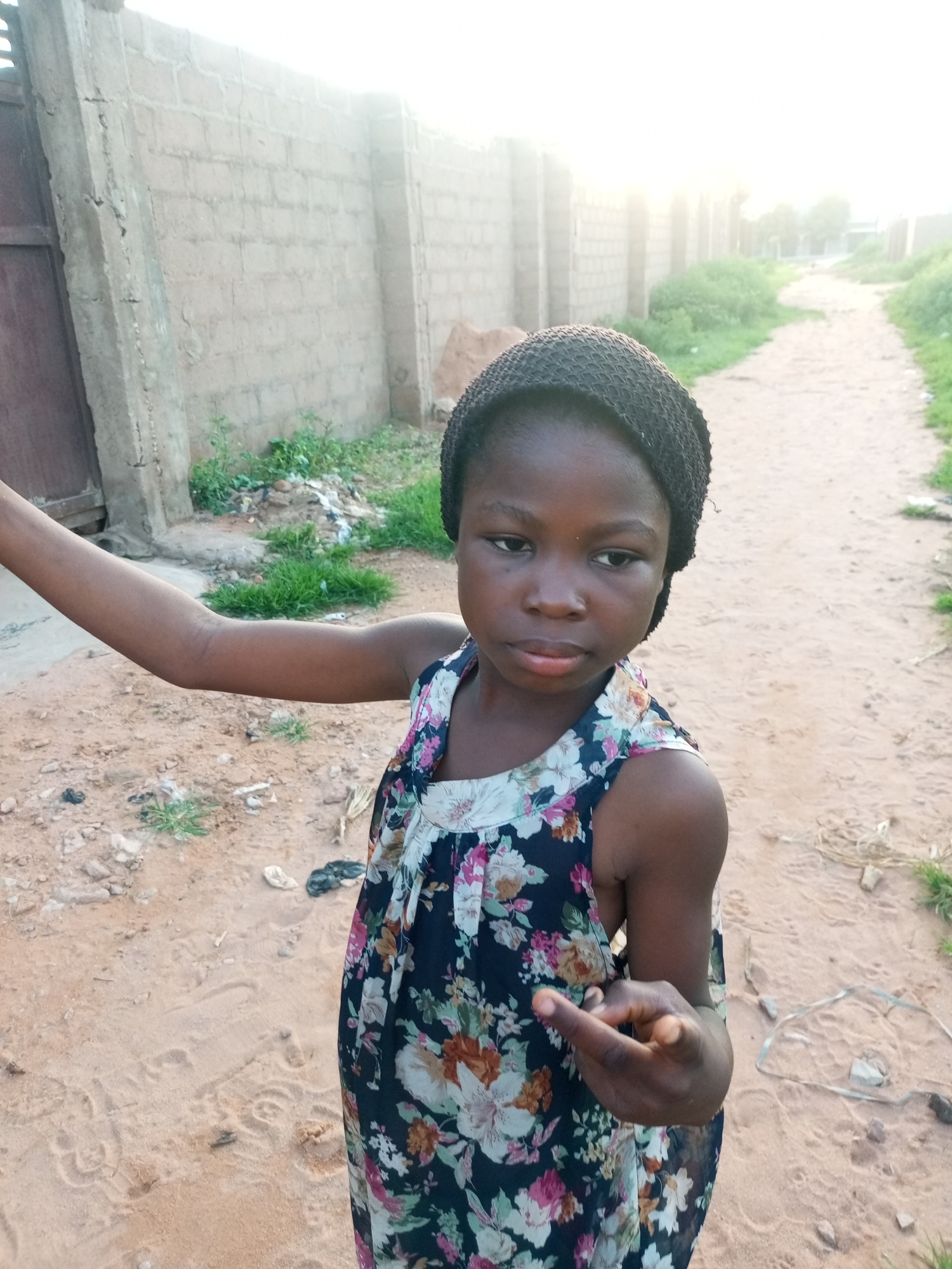 A young Black girl, possibly of Nigerian descent, stands outdoors on a dirt path in a city. She is wearing a dark, sleeveless dress with a floral pattern of white, pink, and orange flowers. On her head, she wears a dark, knitted cap. Her left arm is extended forward and slightly to her left, and her right hand is raised in front of her body, with her index finger pointing.
The background shows a rough, dirt path stretching into the distance, bordered by sparse green vegetation and a low, beige brick wall. To the left of the girl, a dark brown metal gate is visible, adjacent to a grey concrete pillar. The sky is bright and hazy, suggesting either dawn or dusk. The ground is sandy and uneven, with scattered debris, including some white and dark plastic items, and a coil of wire on the right side of the path. The overall scene depicts an outdoor environment, likely in a residential or less developed area of Tempure, Nigeria.