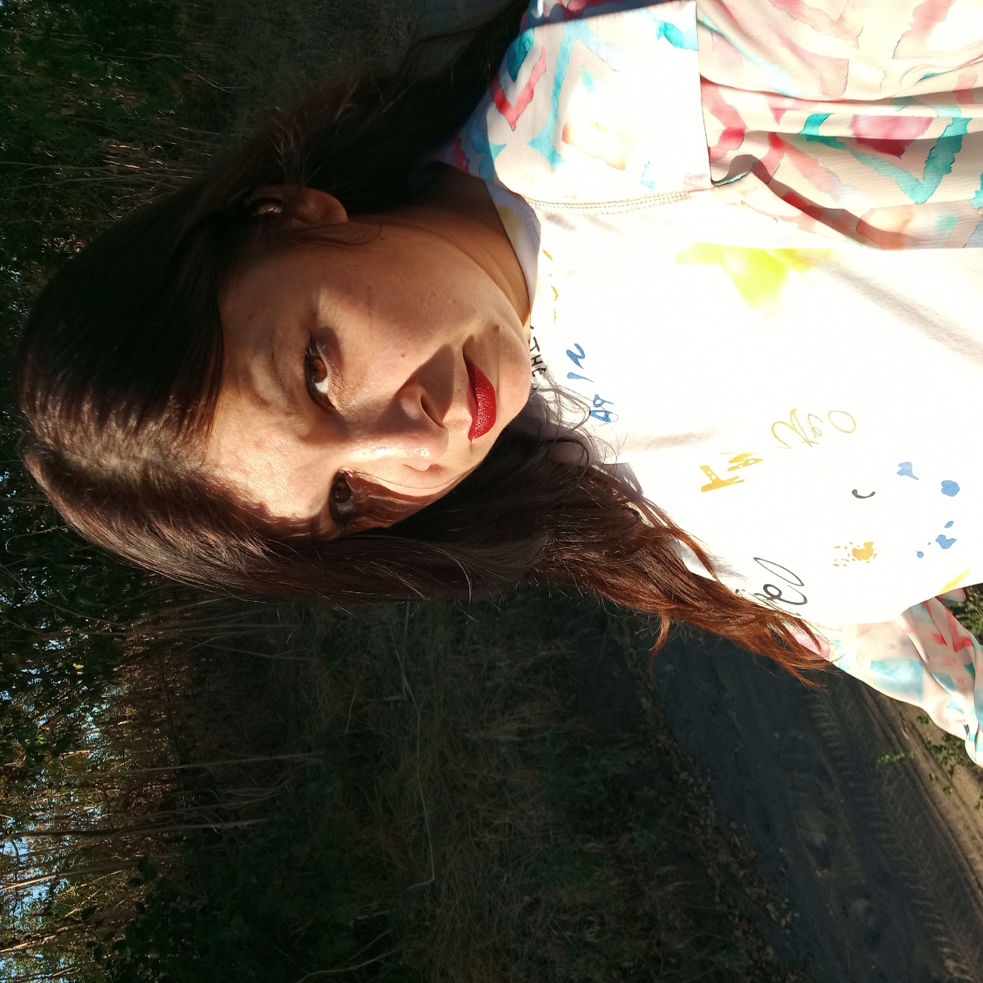 A woman with dark brown hair and brown eyes is outdoors in a natural setting. She is wearing a white t-shirt with colorful abstract designs and a light-colored jacket also with abstract patterns. Her lips are painted red. She is looking directly at the camera with a serious expression. The background consists of tall, thin trees and dry grass, suggesting a rural or park-like environment. The lighting indicates it is daytime, with sunlight casting shadows on the woman's face. No specific details about the city of Klyntsi, Ukraine are visible in the image.