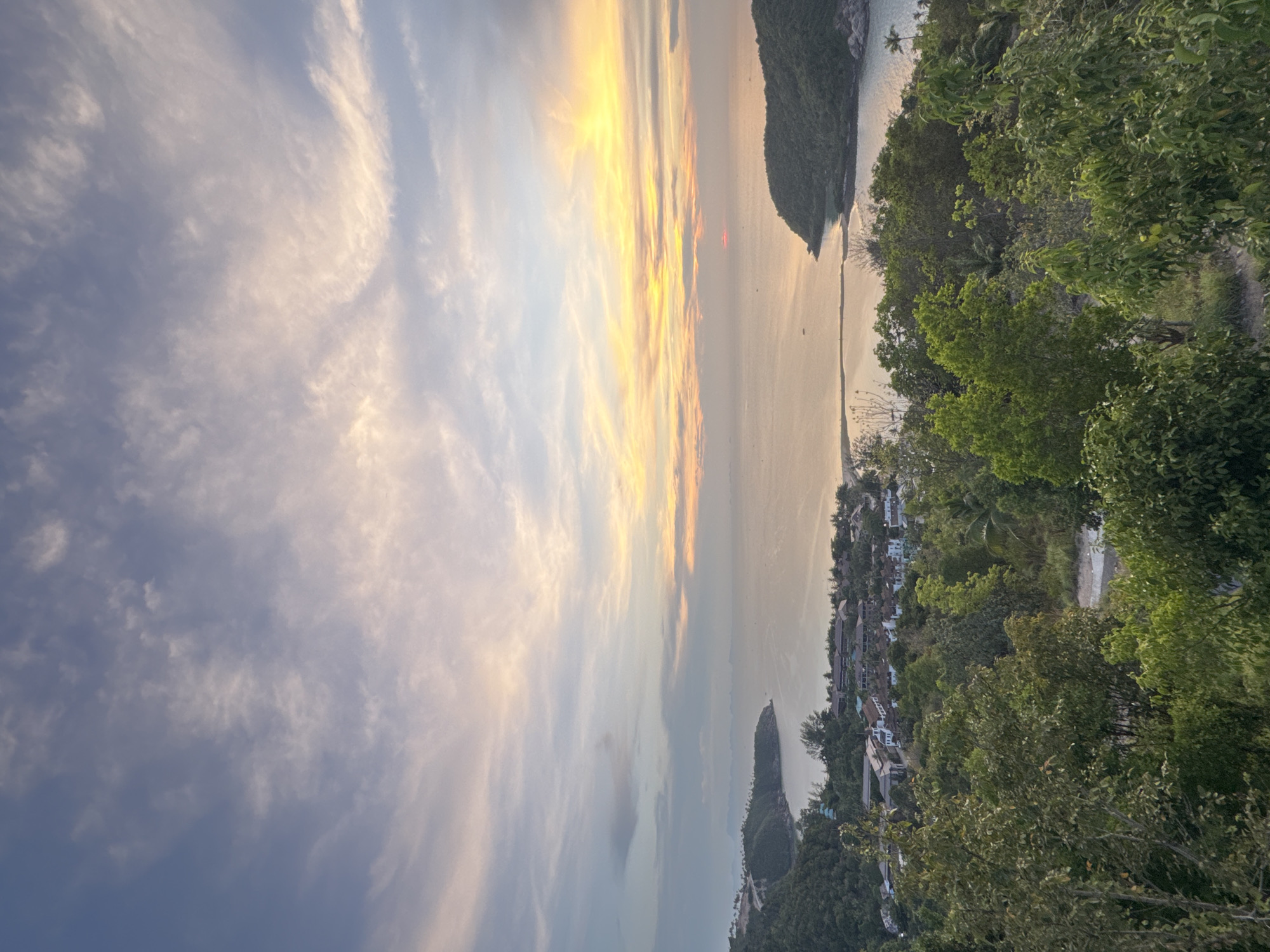 The image shows a sunset scene in Baan Wang Ta Kien, Thailand. The sky is filled with wispy clouds illuminated by the setting sun, creating a gradient of colors from yellows and oranges near the horizon to blues and grays higher up. The sun is reflected on the water's surface. The coastline of the island is partially visible, with green vegetation covering its terrain. A small settlement or town with buildings is nestled along the coast, framed by trees. The foreground is dominated by lush, green vegetation, likely trees, which obscure some of the view but provide a sense of elevation.