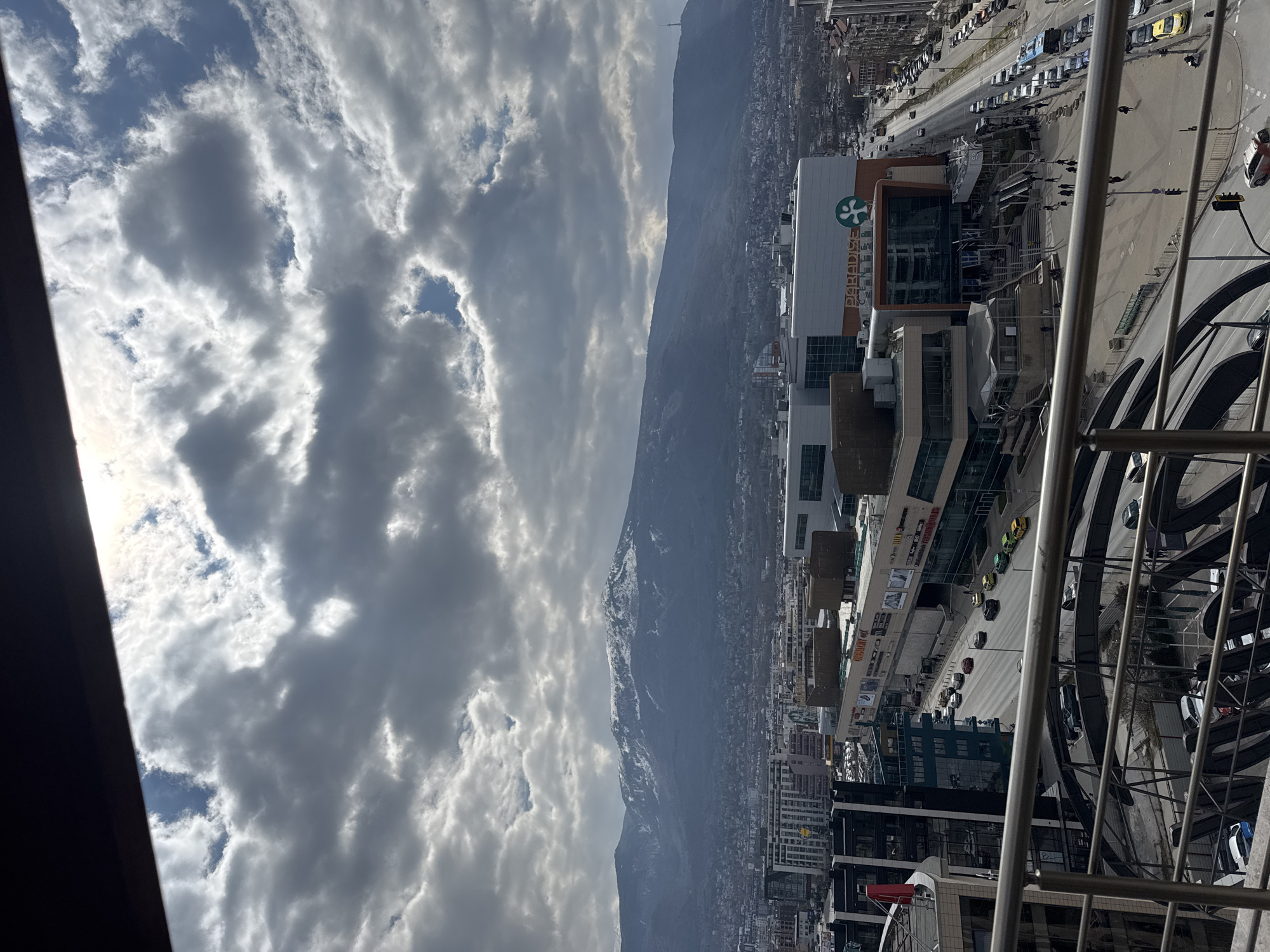 The image provides an elevated, wide-angle view of an urban landscape in Sofia, Bulgaria, centered on the Paradise Center shopping mall. The scene is observed from a high vantage point, with dark metal balcony railings partially visible across the immediate foreground.
The sky is characterized by a significant covering of large, grey cumulus clouds, interspersed with clear patches of blue sky and areas of bright sunlight. In the background, the snow-capped Vitosha Mountain range dominates the horizon, rising above the sprawling city.
In the mid-ground, the multi-story Paradise Center shopping mall is the most prominent structure, identifiable by its name "PARADISE CENTER" and a green tree-like logo on its facade. The mall exhibits modern architecture with extensive glass panels and light-toned exterior surfaces. Its main entrance features multiple outdoor escalators leading to an upper level. Surrounding the mall are various other multi-story commercial and residential buildings.
Below the mall and in the immediate foreground, a complex network of roads is active with vehicular traffic, including numerous cars, several yellow taxis, and a blue public bus. Pedestrians are visible traversing sidewalks, utilizing crosswalks, and walking within the large open plaza area directly in front of the mall's entrance. Streetlights and traffic signals are also present along the roadways. The overall scene depicts a bustling urban environment under dynamic cloud cover.