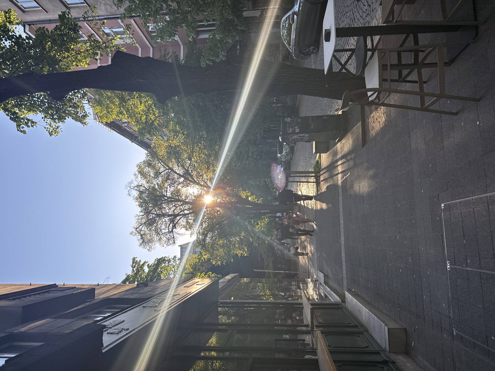 The image shows a street scene in Sofia, Bulgaria, with pedestrians walking on a sidewalk. The sun shines brightly through the trees, creating lens flare and casting shadows. On the right, there is outdoor seating of a cafe or restaurant, with tables and chairs. On the left is a building with a glass facade. Cars are parked along the street.