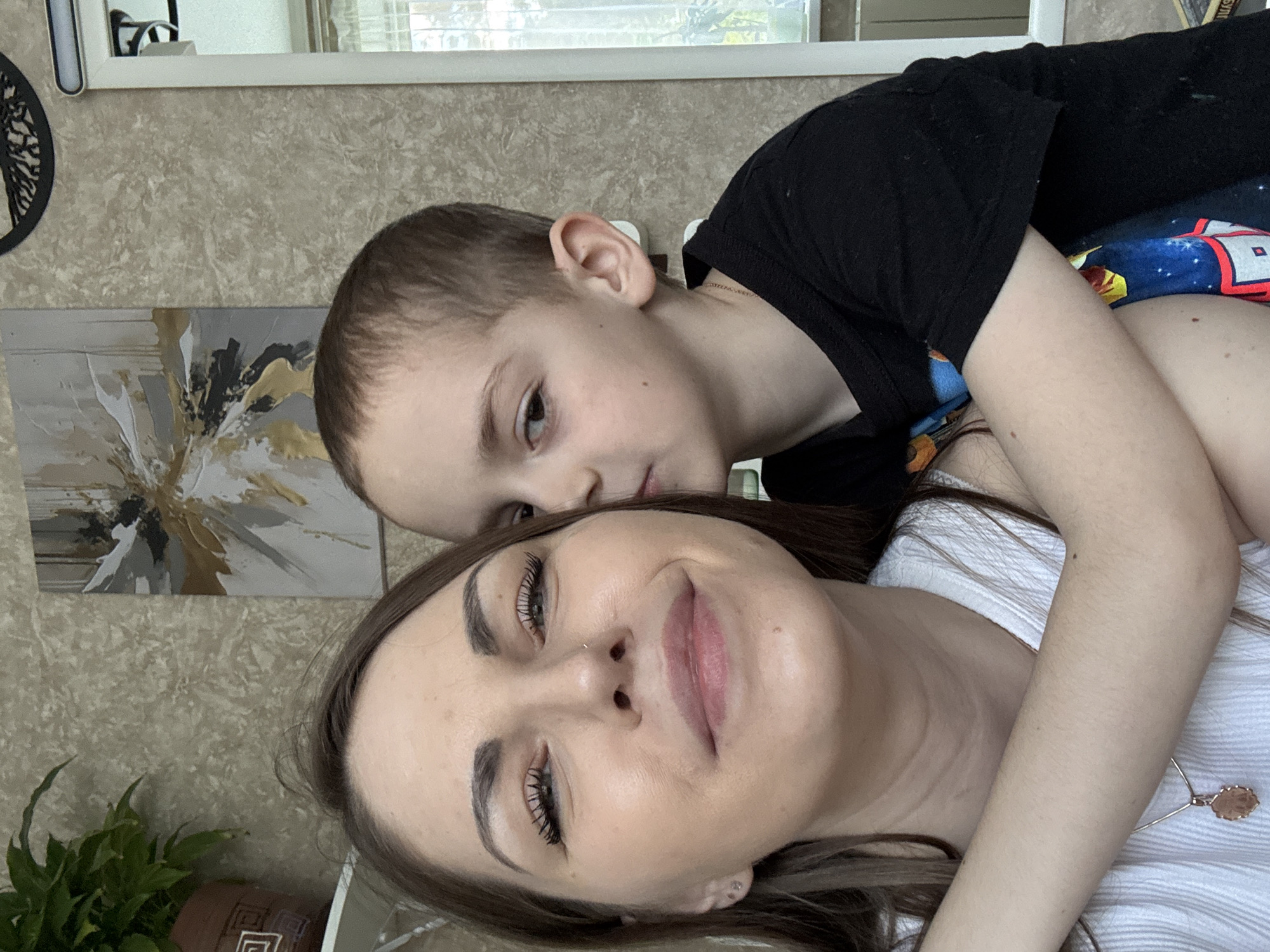 A woman and a child are closely posed together. The woman has fair skin, dark eyebrows, long eyelashes, and a nose piercing. She is wearing a white, collared shirt and a necklace with a circular pendant. The child, a young boy, has short brown hair. He is wearing a black shirt with a colorful graphic design. The boy's arm is draped across the woman's chest.
The background features a patterned wallpaper, a painting with gold and grey accents, and a green potted plant. A white-framed mirror is visible in the upper left corner. The location within Раздолье, Russia, is not discernible from the image.