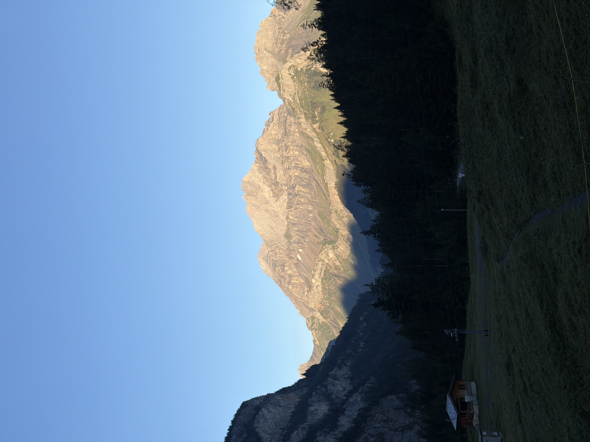 A wide-angle, vertical, natural light photograph depicts a mountain valley landscape under a clear blue sky at what appears to be sunrise or sunset. In the foreground, a dark green, gently sloped meadow is visible, with a small, wooden structure with a brown roof situated on the left side. A narrow, light-colored path winds through the grass. A dark, dense forest of coniferous trees forms a line across the middle ground, separating the meadow from the towering, rugged mountains beyond. The mountains are illuminated by warm, golden light on their peaks and upper slopes, casting deep shadows below. The rugged terrain of the mountains is characterized by exposed rock faces and sparse vegetation. The sky is a gradient of light blue, free of clouds. There are no people or animals visible in the image. The scene appears to be a remote, natural environment. This location is likely in the vicinity of Kandersteg, Switzerland, known for its alpine scenery and hiking trails.