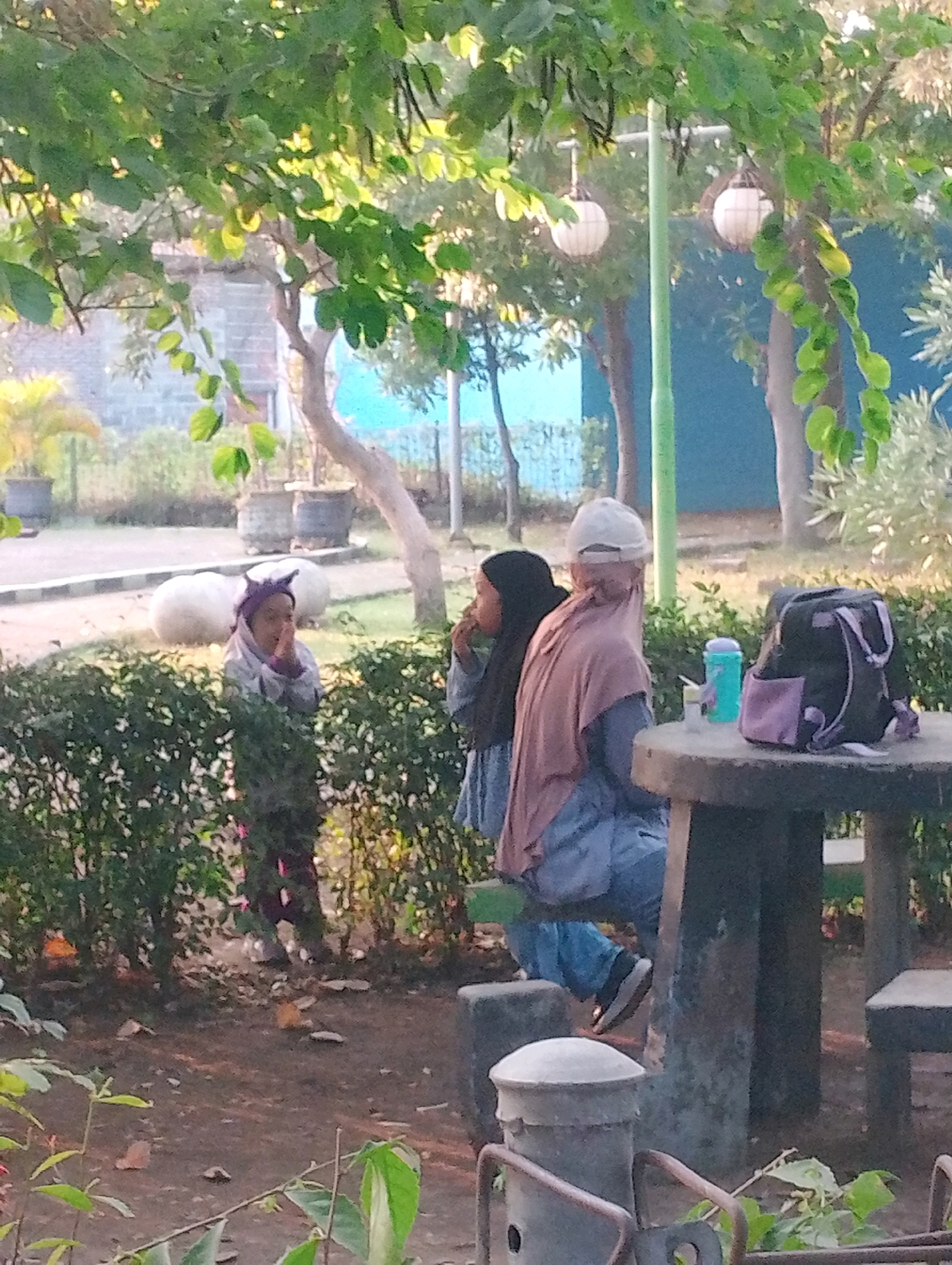 The image depicts an outdoor scene in Probolinggo, Indonesia, likely a park or public recreational area. In the foreground, a circular stone table with two stone benches is visible. On the table, a black backpack with purple accents, a teal water bottle, and a small white cup are placed.
To the right of the table, a person wearing a light-colored hijab and a light brown shawl sits on a bench, facing away from the camera. They are wearing blue pants and black shoes. To the left of this person, another individual wearing a black hijab is partially visible, also seated.
In the middle ground, a young child wearing a purple head covering and a grey jacket stands behind a hedge, with their hands clasped in front of their face. Two large white round objects are visible behind the child, possibly decorative elements or seating.
The background features lush green trees and bushes, with sunlight filtering through the leaves. In the distance, a blue wall is visible, along with two white spherical lamps hanging from poles. The ground is covered in dirt and scattered leaves. The overall impression is a tranquil, everyday scene in a public space in Probolinggo.