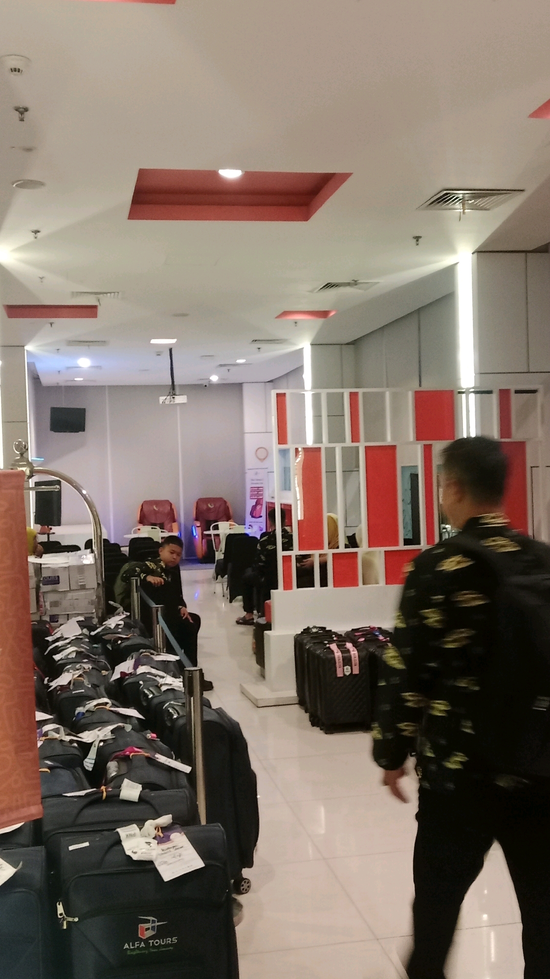 The image depicts an indoor waiting area, likely at an airport or train station, in Tangerang, Indonesia. Several pieces of black luggage are arranged in rows, some with tags attached. In the foreground, a person in a black jacket with a floral pattern is walking away from the camera. In the middle ground, a young person in a black outfit is seated, looking down. Behind them, two massage chairs are visible, and further back, a projector is mounted on the ceiling. The environment features a white tiled floor, white walls, and a white ceiling with recessed red lighting fixtures. A decorative partition with red and white vertical elements is also present.