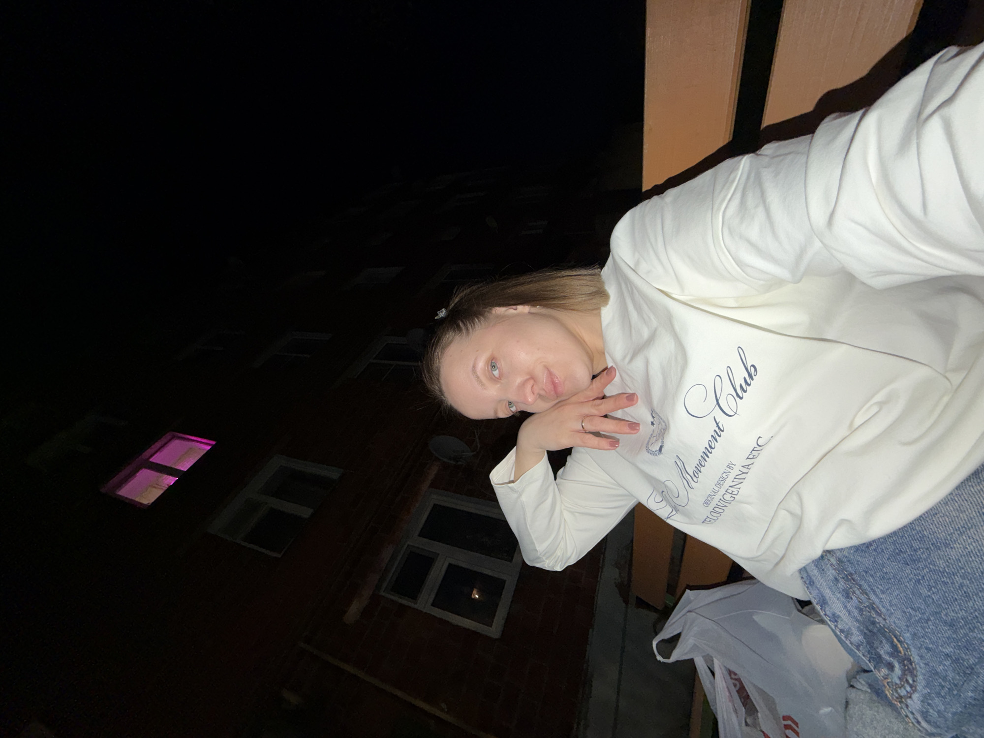 A woman poses for a selfie on a balcony at night. She is wearing a white sweatshirt with the words "Movement Club" and blue jeans. Behind her is a brick building with lit windows. The building is located in Reshetnikovo, Russia. The sky is dark. A partially visible white plastic bag is in the lower right.
